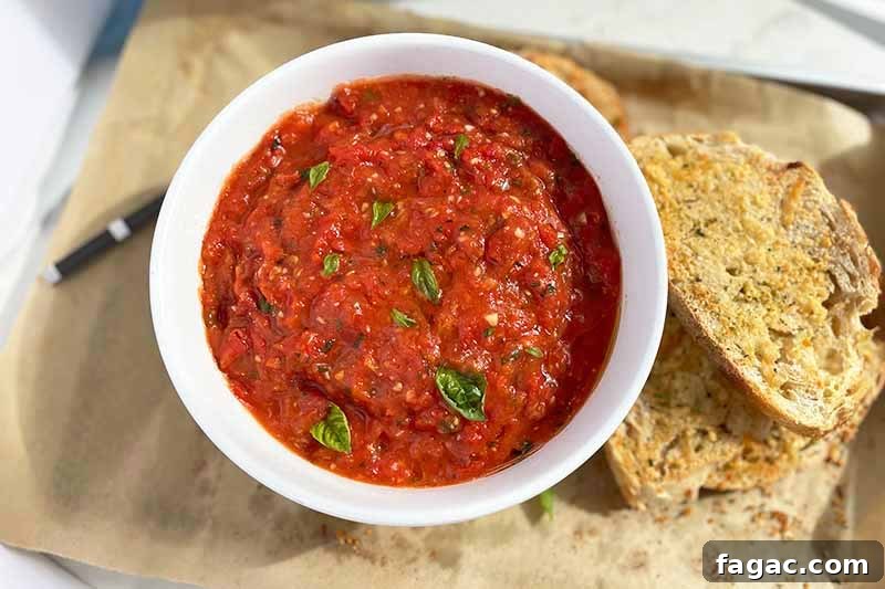 Oven-Roasted Tomato Sauce Recipe for Rich Homemade Flavor 12 Horizontal image of a white bowl with a red sauce next to slices of garlic bread on a parchment paper lined baking sheet.
