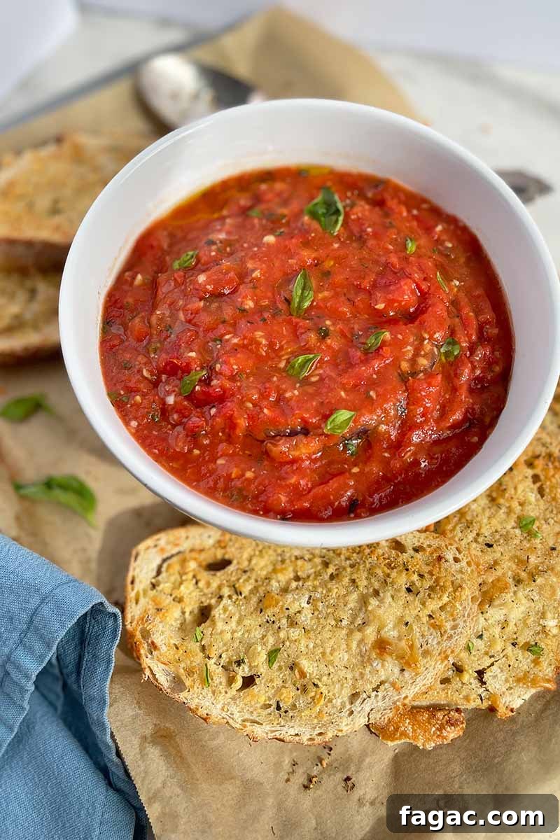 Oven-Roasted Tomato Sauce Recipe for Rich Homemade Flavor 4 Vertical image of a white bowl with a thick red liquid on a baking sheet with garlic bread.