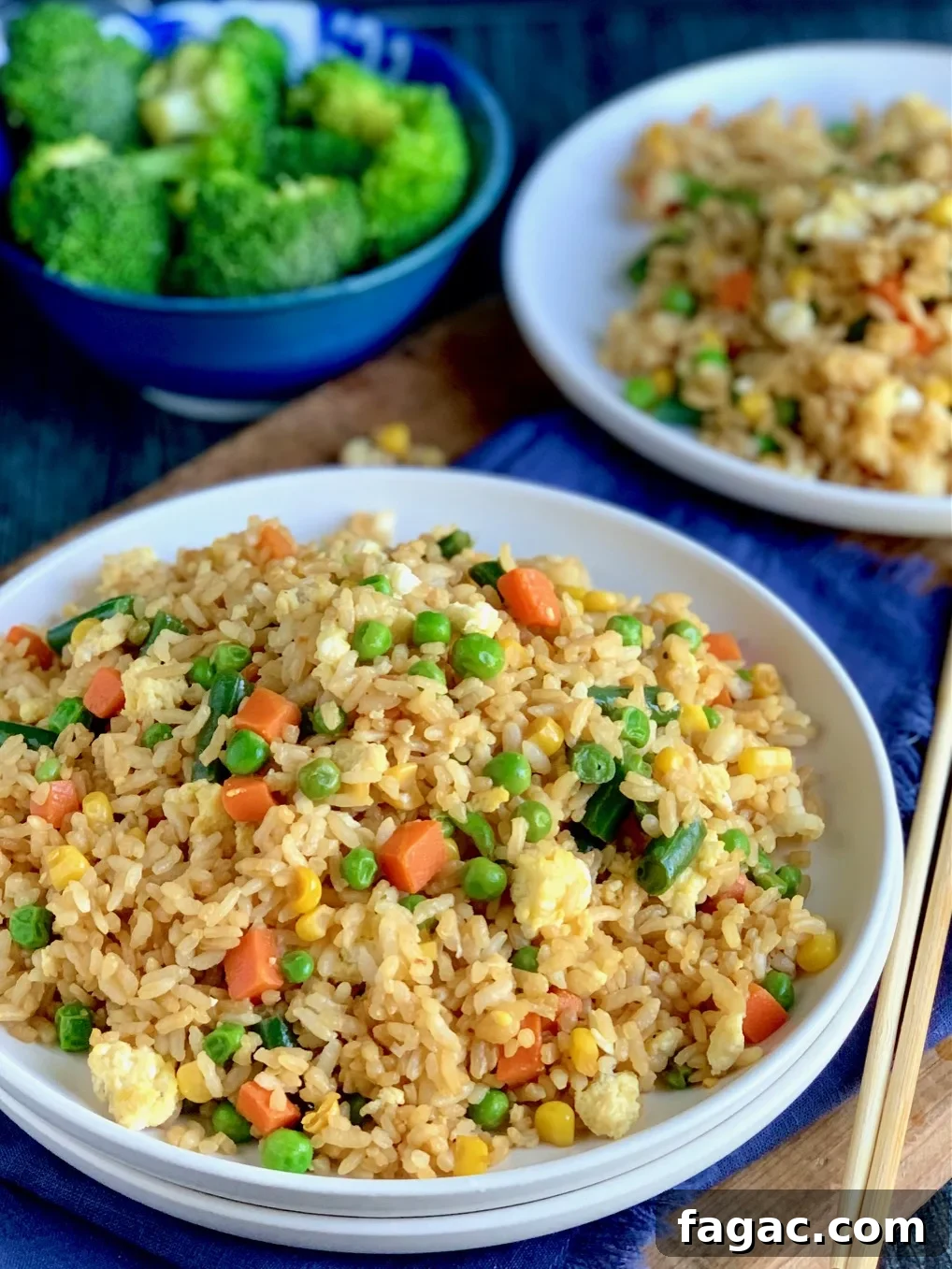 A large white plate piled with golden brown rice with small cubed carrots, peas, corn, and green beans next to chopsticks and a bowl of broccoli.