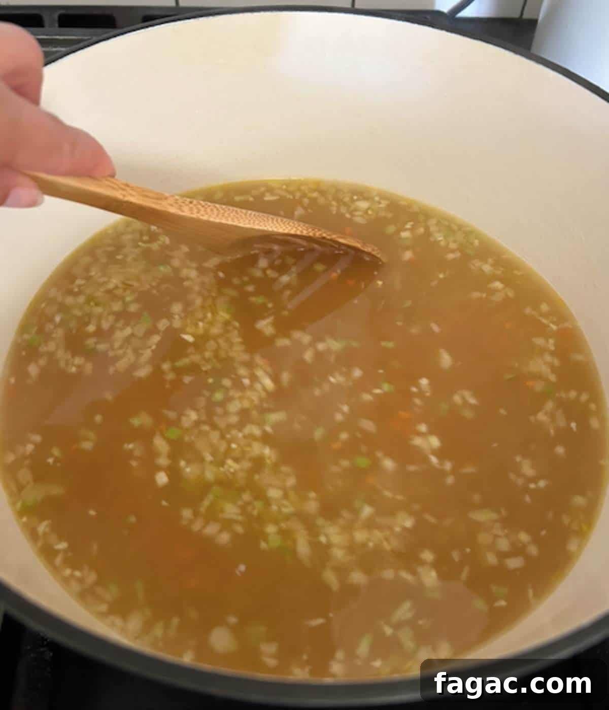 Hand stirring pastina into soup with wooden spoon.