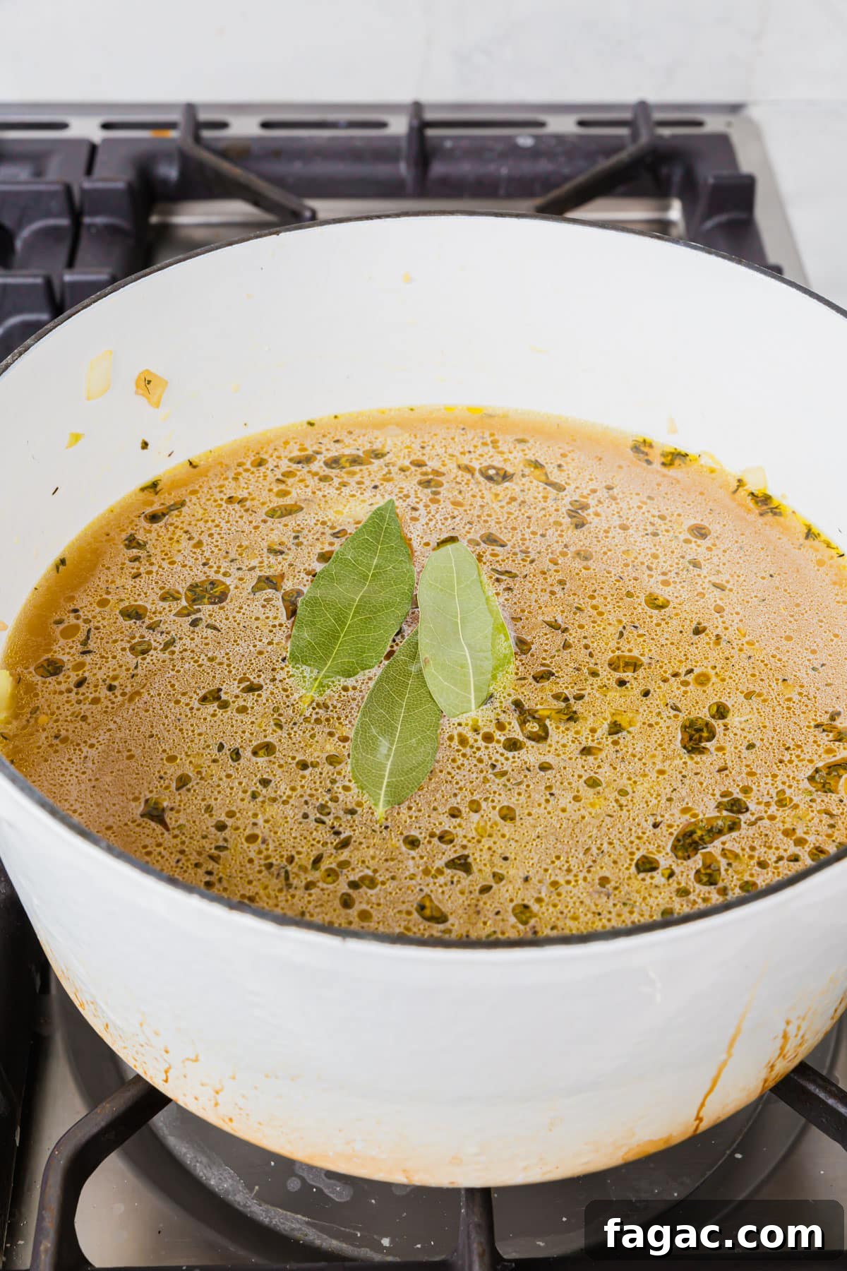 Comforting Gluten-Free Chicken Noodle Soup Recipe 7 A white dutch oven of chicken broth, herbs, diced onion and dried bay leaves.