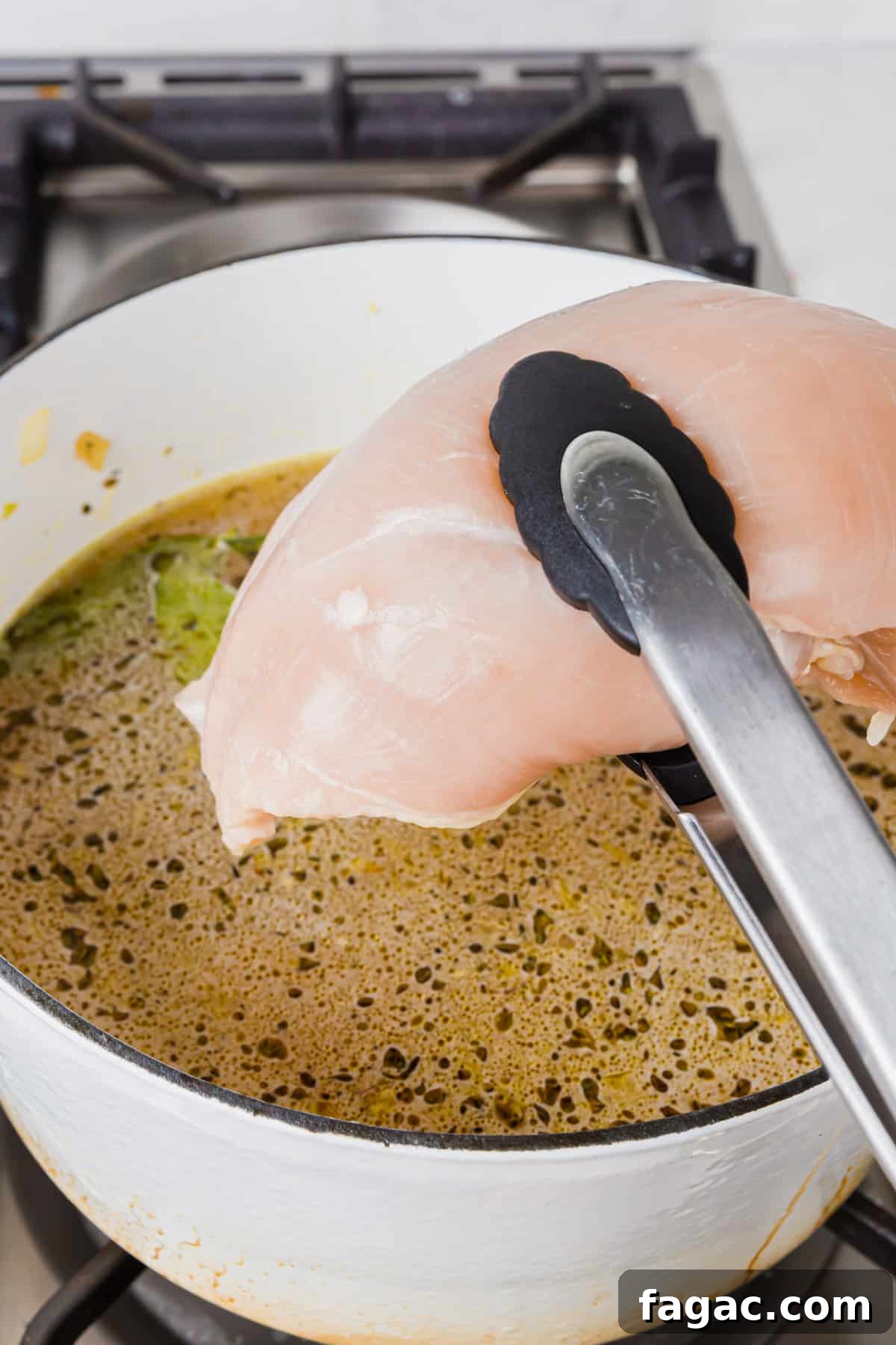 Comforting Gluten-Free Chicken Noodle Soup Recipe 8 A pair of tongs lowering a raw chicken breast into a dutch oven of chicken broth, herbs, bay leaves, and minced garlic.