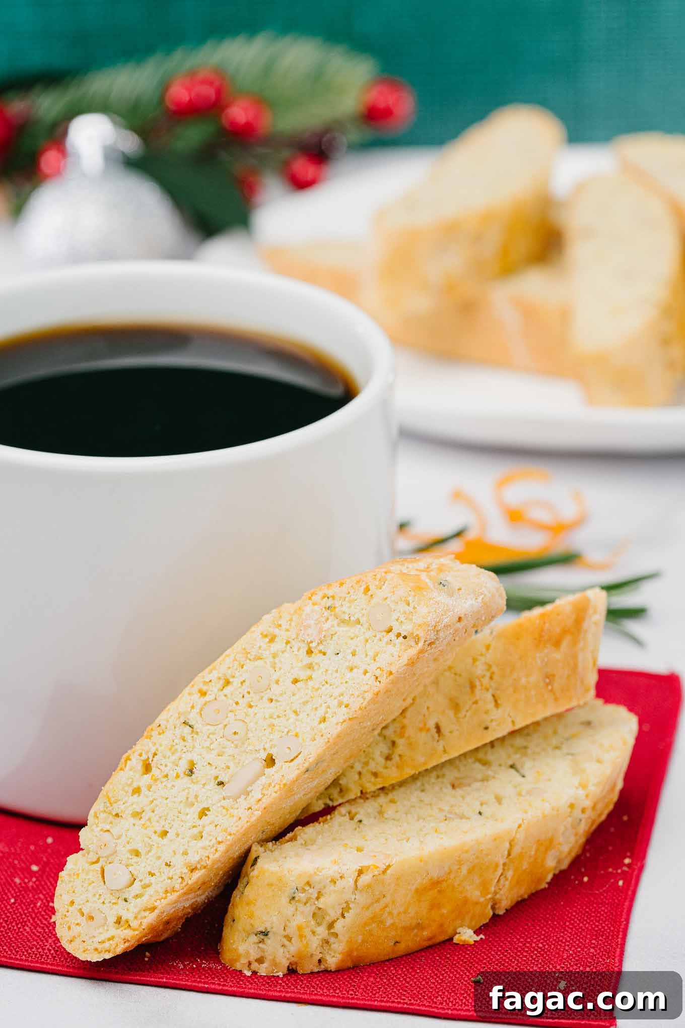Buttery Orange Biscotti: Classic and Almond Variations 2 Buttery biscotti with orange, pine nuts, and rosemary on a red napkin next to a mug of coffee, more biscotti and Christmas table decor in the background.