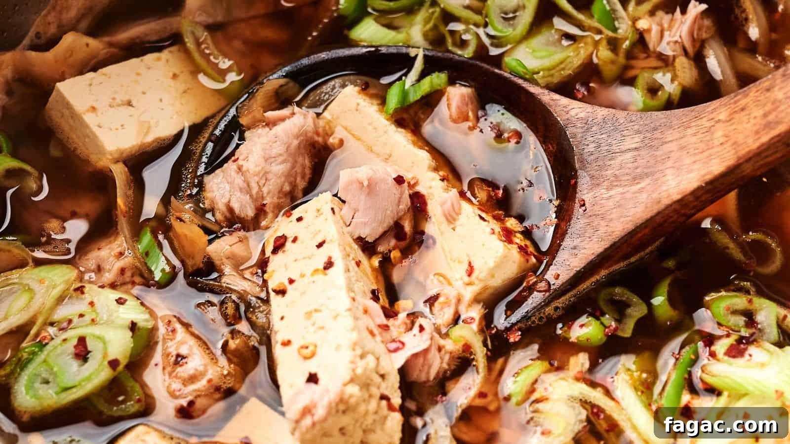 Close-up of a wooden spoon in a vibrant kimchi stew with tofu and pork, garnished with sliced green onions and red pepper flakes.