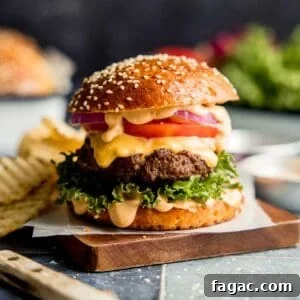 Hamburger topped with sauce, tomato, onion, and lettuce on wood cutting board.
