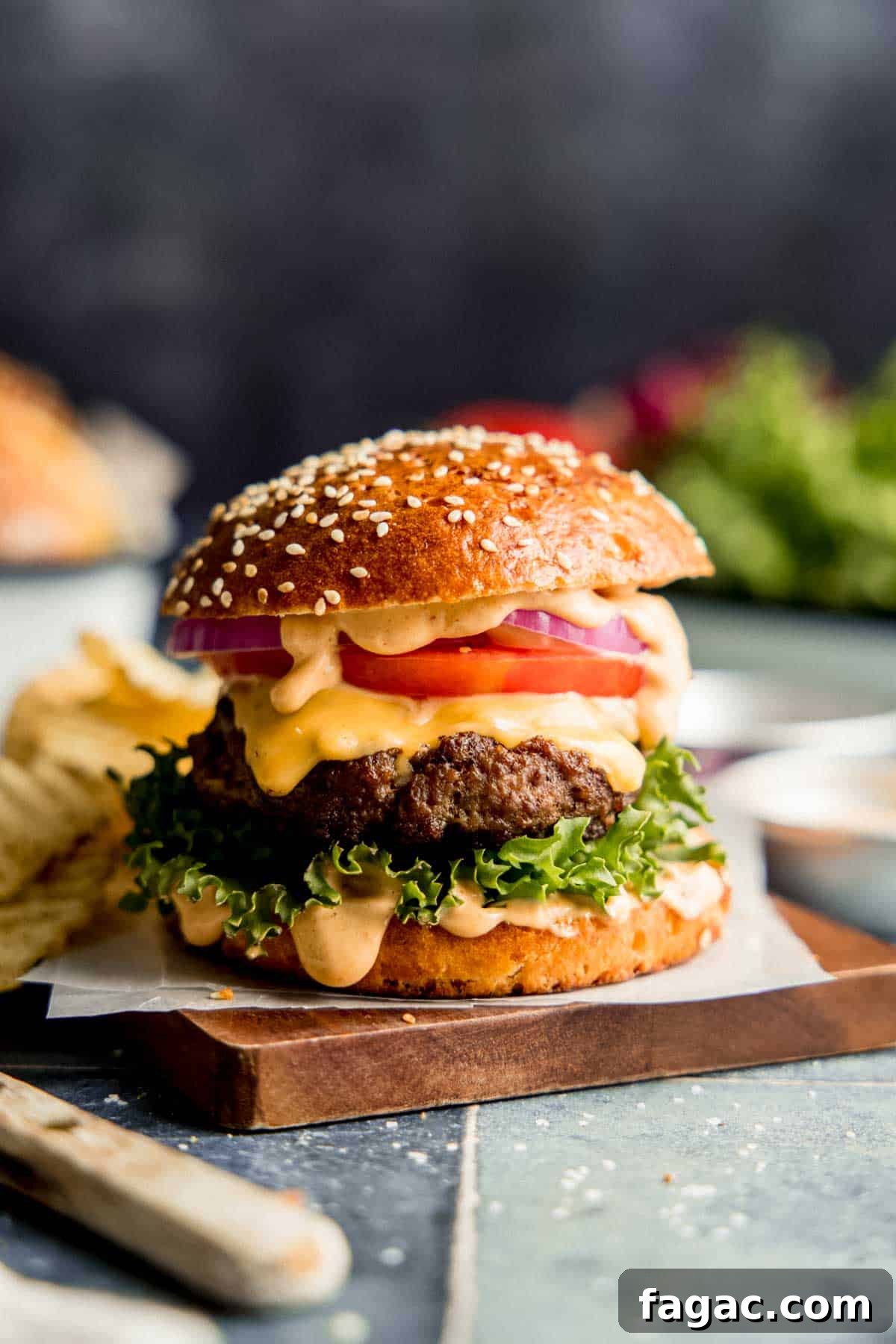 Burger with lettuce, tomato, onion, and sauce on a sesame seed bun, resting on wooden board.