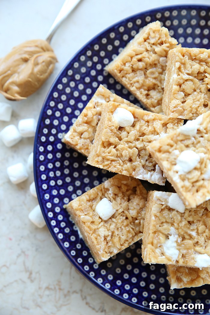 A large dark blue round Polish pottery plate with white polka dots with square peanut butter Rice Krispie treats stacked on each other. On the left of the plate is a spoon with creamy peanut butter on it and several mini marshmallows.