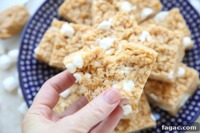 A partial torn peanut butter Rice Krispie treat held above a round dark blue polka dotted Polish potter plate with cut Rice Krispie treats stacked on each other. Faded in the upper left corner is a small spoon with peanut butter with several mini marshmallows below it and along the side of the plate.
