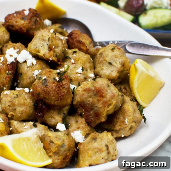 A bowl of Greek air fryer chicken meatballs with slices of lemon and a spoon in - there's also a Greek salad in the background
