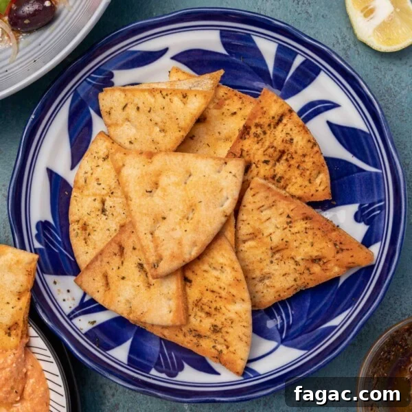 Homemade pita chips in a blue floral bowl with feta dip, lemon halves, Greek salad and salt just visible at the sides, all on a mottled blue background.