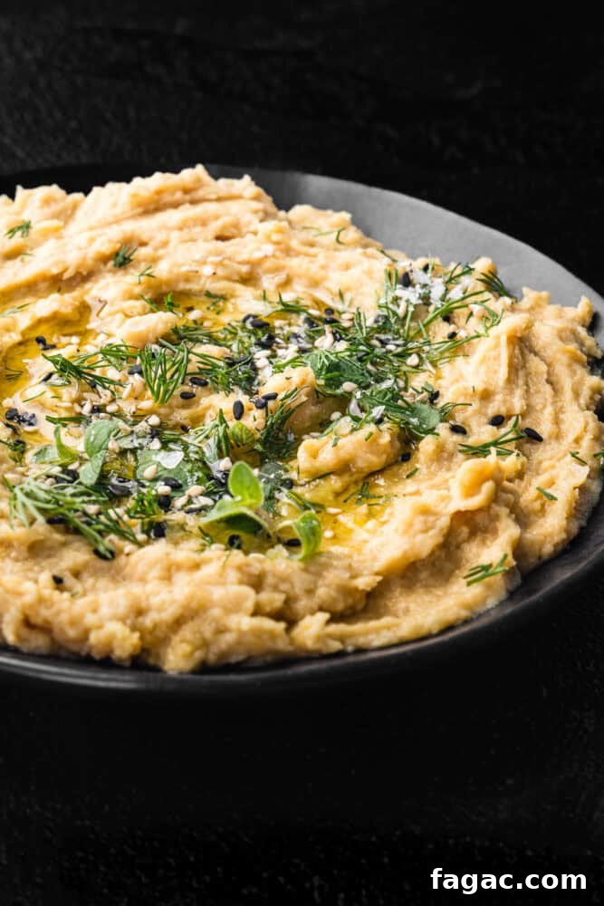 Hummus without tahini topped with fresh dill, sesame seeds, olive oil, and flaky salt in a black bowl on a dark background, close-up angled shot