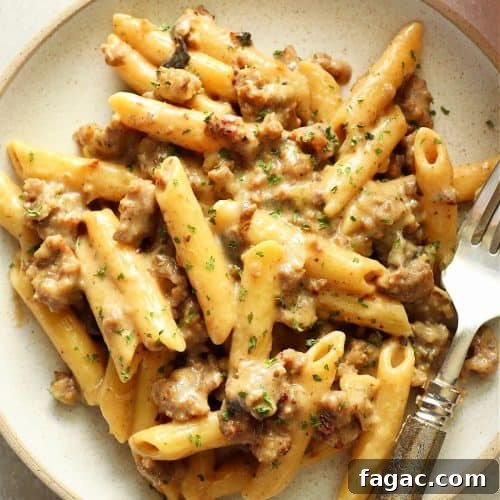 Square image of penne pasta with sausage and creamy sauce on a speckled plate with fork.