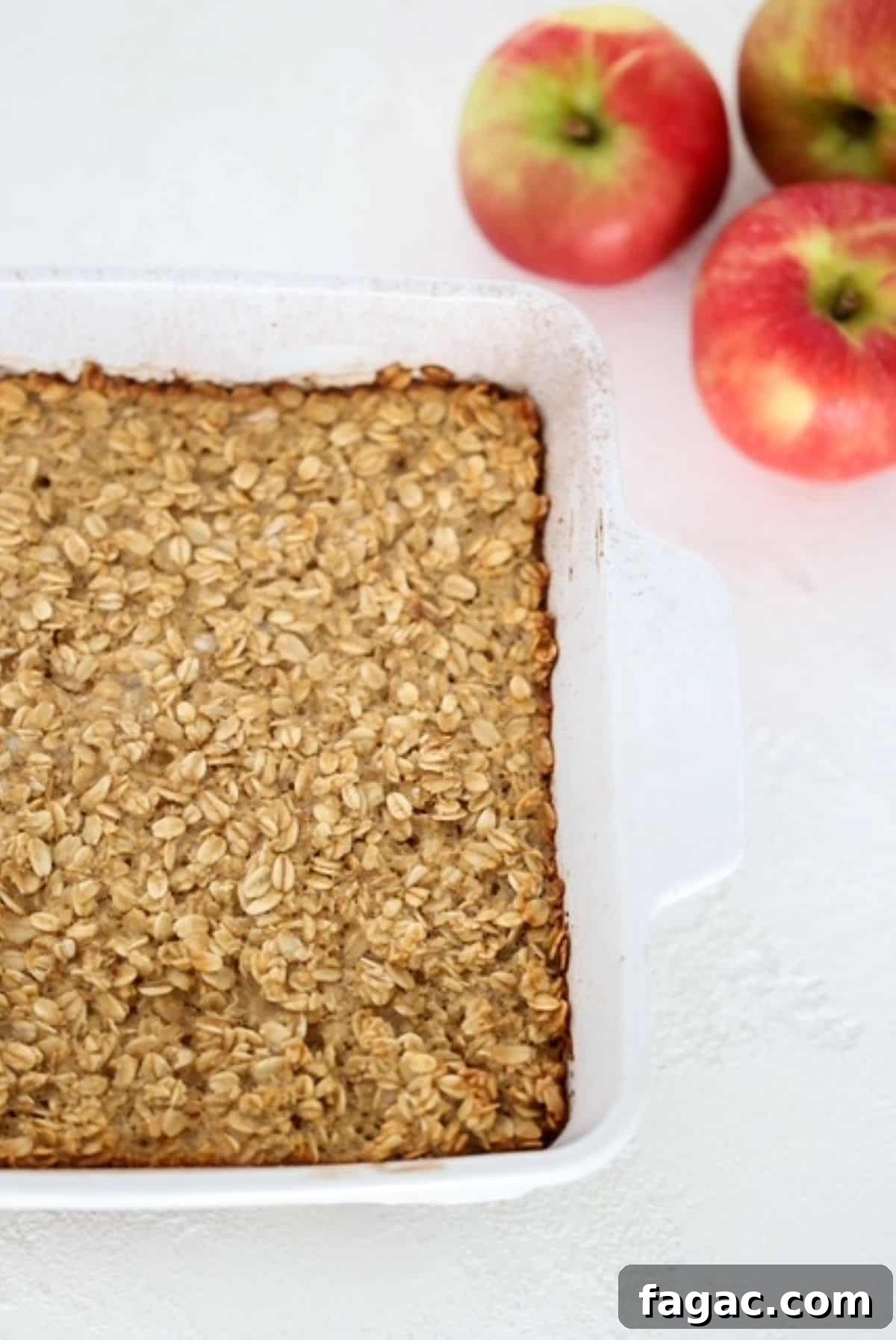 Apple Cinnamon Baked Oatmeal Recipe for Cozy Breakfasts 5 Apple Cinnamon Baked Oatmeal in a white baking dish with three apples to the top right of the baked oatmeal.