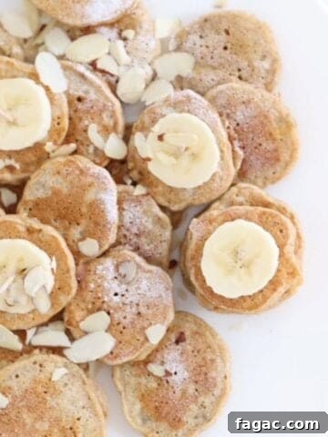 Mini banana pancakes dusted with powdered sugar, topped with sliced banana and almond slices on a white plate.