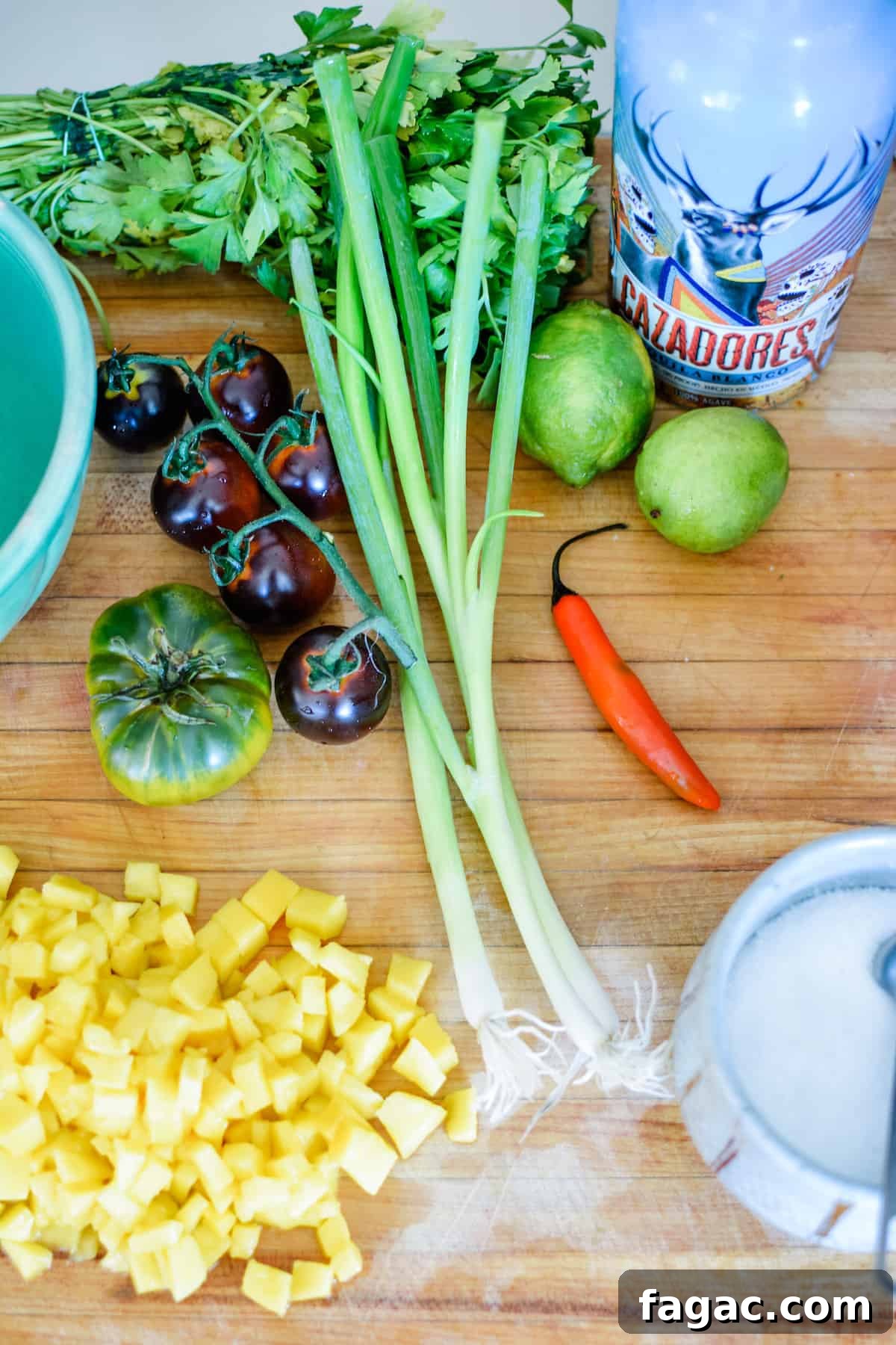 A bottle of tequila, two limes, a red serrano Chile, a bowl of salt, three scallions, several tomatoes, and mango all on a wood cutting board. 