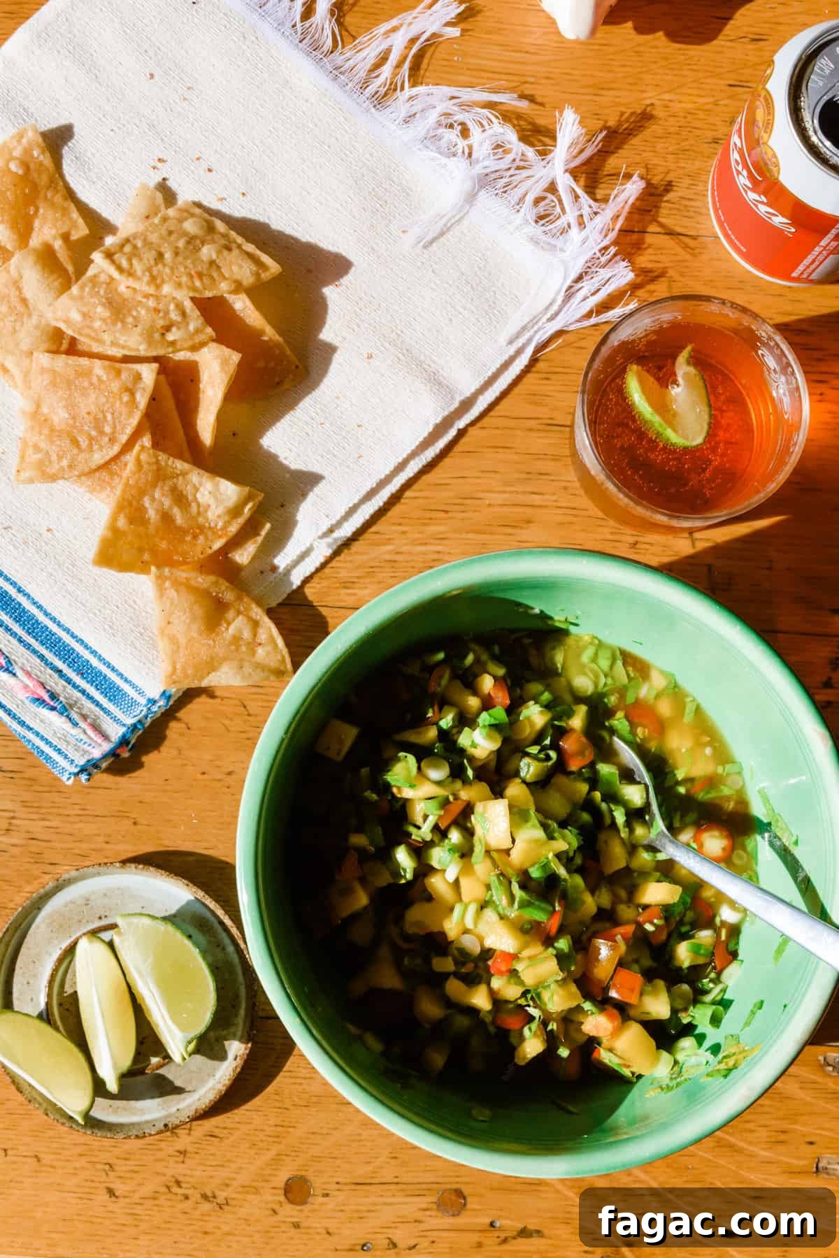 A green bowl with mango margarita salsa sitting on a wooden table with lime wedges, tortilla chips, and a beer on the side.