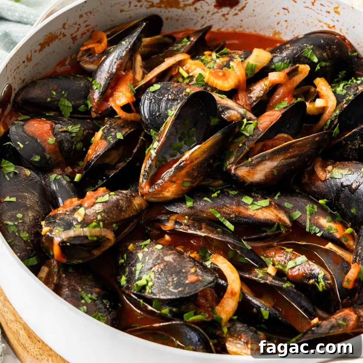 Frying pan with steamed mussels in tomato sauce, sprinkled with a parsley garnish, and some pieces of bread around the edge.