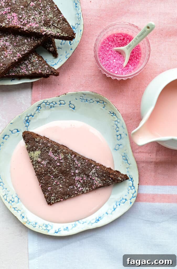 A triangular piece of chocolate concrete dessert with pink sprinkles sits on a floral plate, surrounded by pink custard. Nearby, a small bowl of pink sprinkles and a pitcher of vibrant pink sauce rest on a pink and white cloth.
