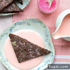 A triangular piece of chocolate concrete dessert with pink sprinkles sits on a floral plate, surrounded by pink custard. Nearby, a small bowl of pink sprinkles and a pitcher of vibrant pink sauce rest on a pink and white cloth.