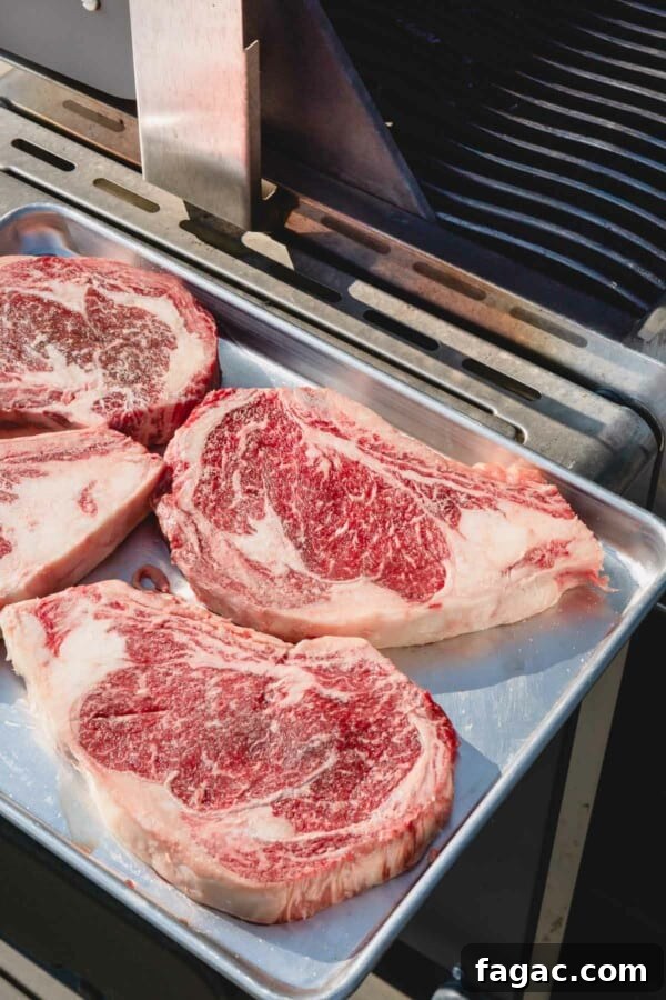 How to Grill a Tender, Flavorful Steak Every Time 5 4 raw ribeye steaks on a baking sheet by a grill.