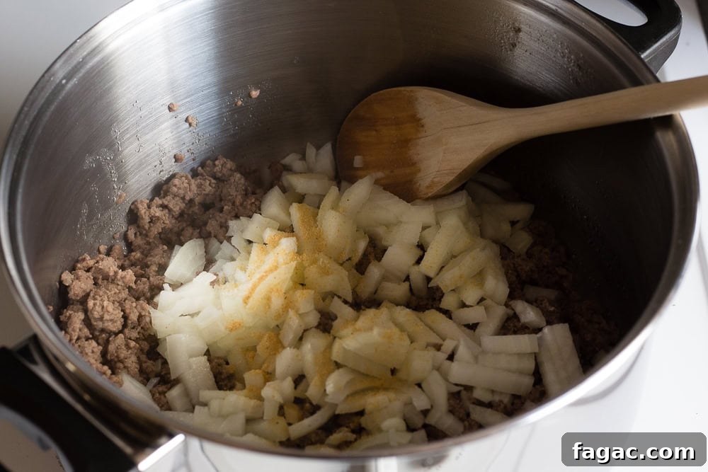 Ground beef and onions in a large silver pot with spices. A wooden spoon rests in the pot as well.