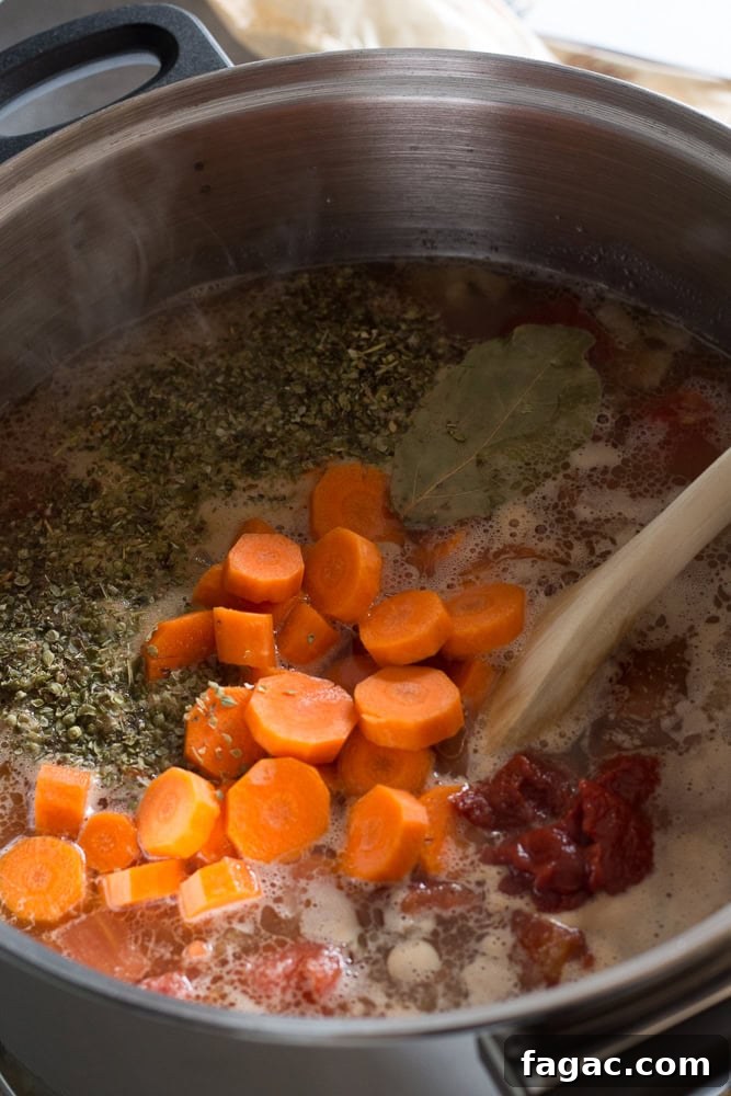 A large pot with soup ingredients in it, including herbs, sliced carrots, tomato paste, and a bay leaf. A wooden spoon rests in the pot as well.
