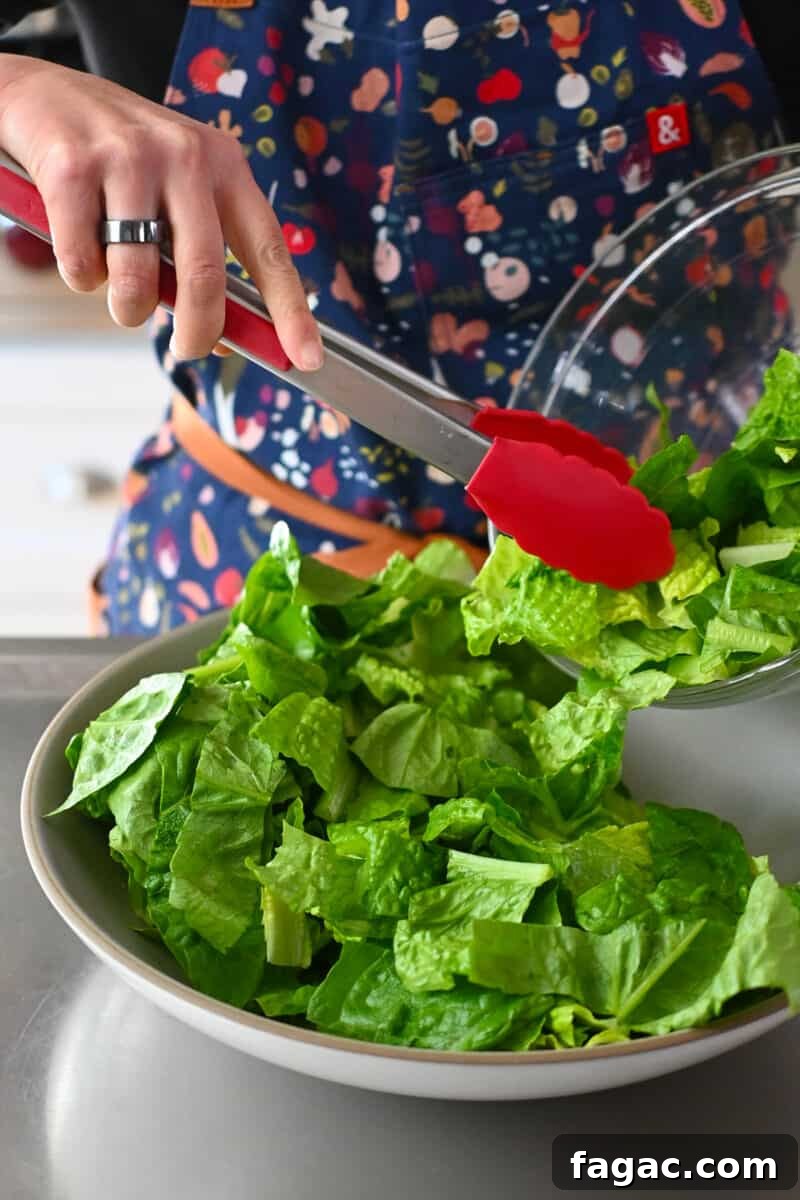 Using red tongs to place chopped romaine lettuce in a shallow salad serving bowl.