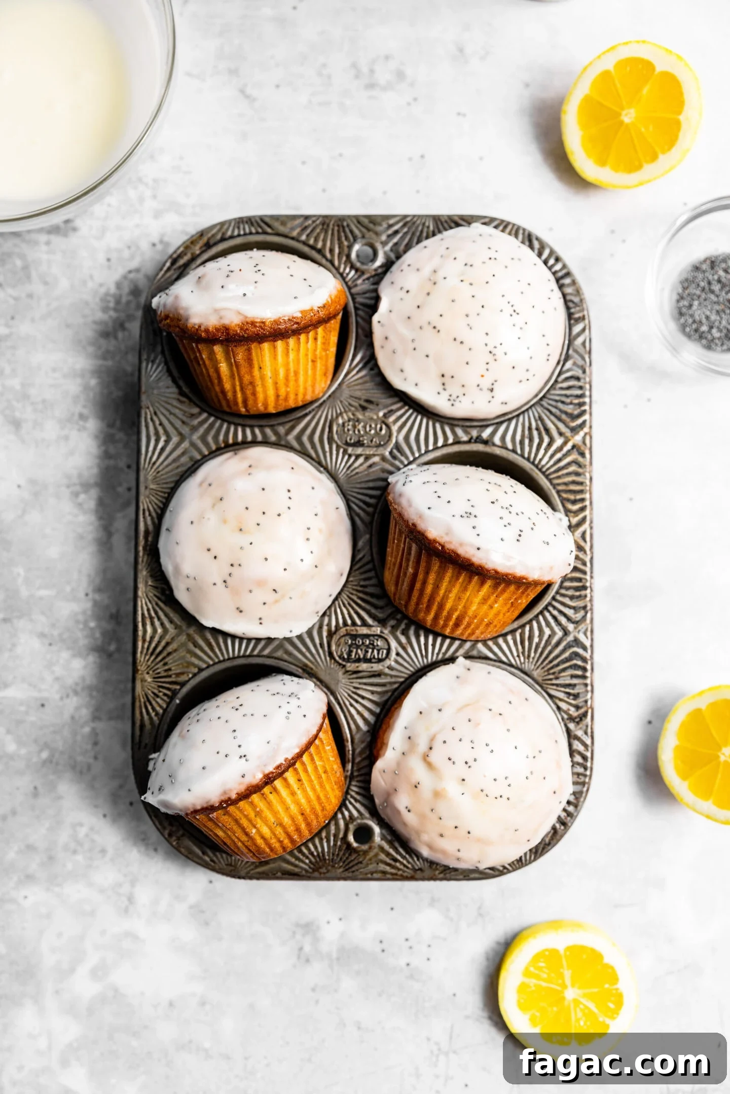 Glazed lemon poppy seed muffins inside of a muffin pan next to lemon slices and small bowl of poppy seeds