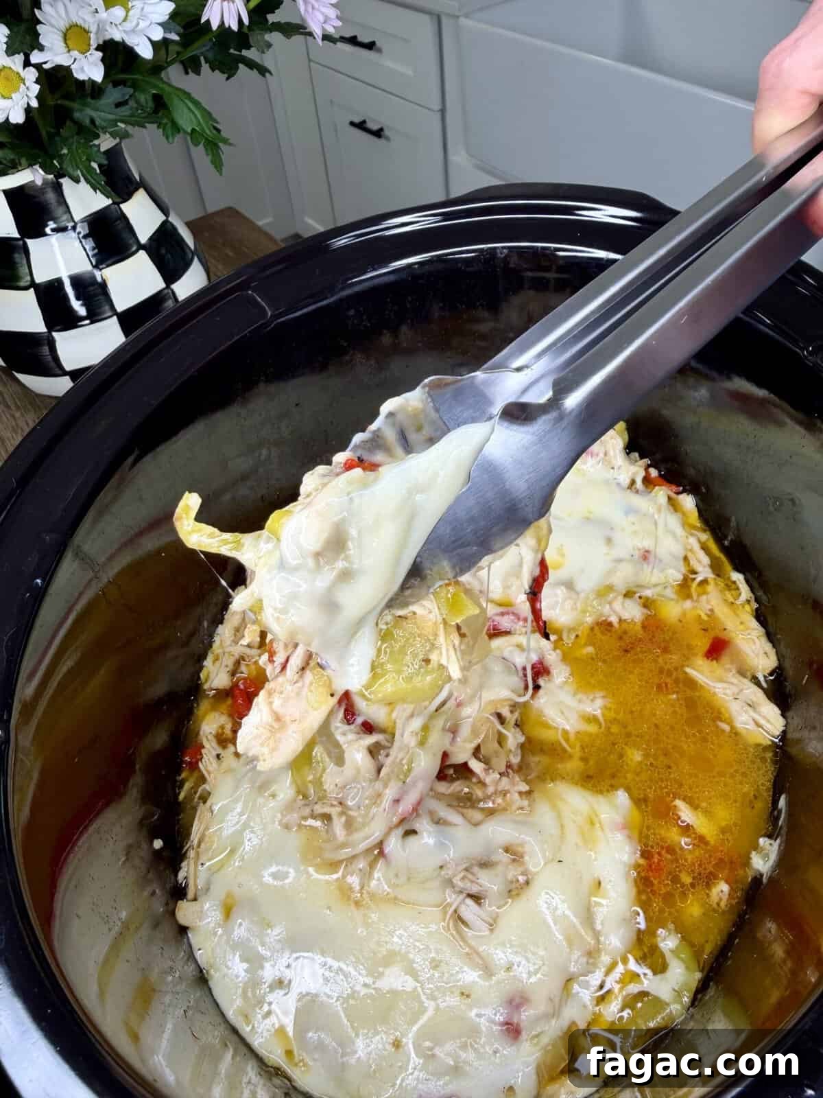 Crockpot Chicken Philly Cheesesteak Recipe for Slow-Cooked Sandwiches 11 A close-up of an Easy Crockpot Chicken Philly Cheesesteak shows melted cheese, shredded chicken, and veggies in a slow cooker. A hand lifts a portion with tongs while a black-and-white checkered vase with flowers decorates the counter nearby.