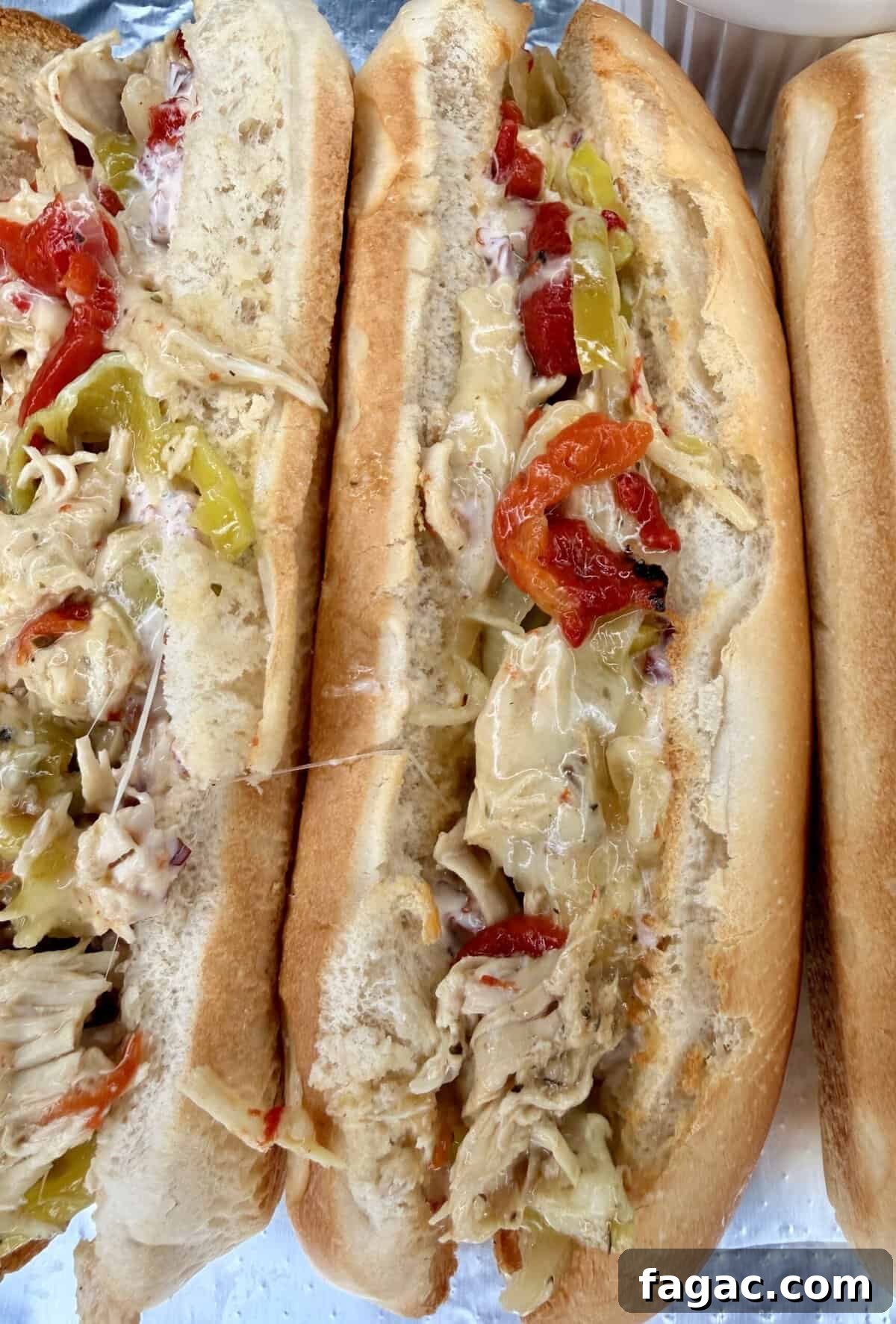 Crockpot Chicken Philly Cheesesteak Recipe for Slow-Cooked Sandwiches 12 Three toasted hoagie rolls filled with Easy Crockpot Chicken Philly Cheesesteak—shredded chicken, melted cheese, sliced red peppers, and yellow pepper rings—are arranged closely together on a silver tray.