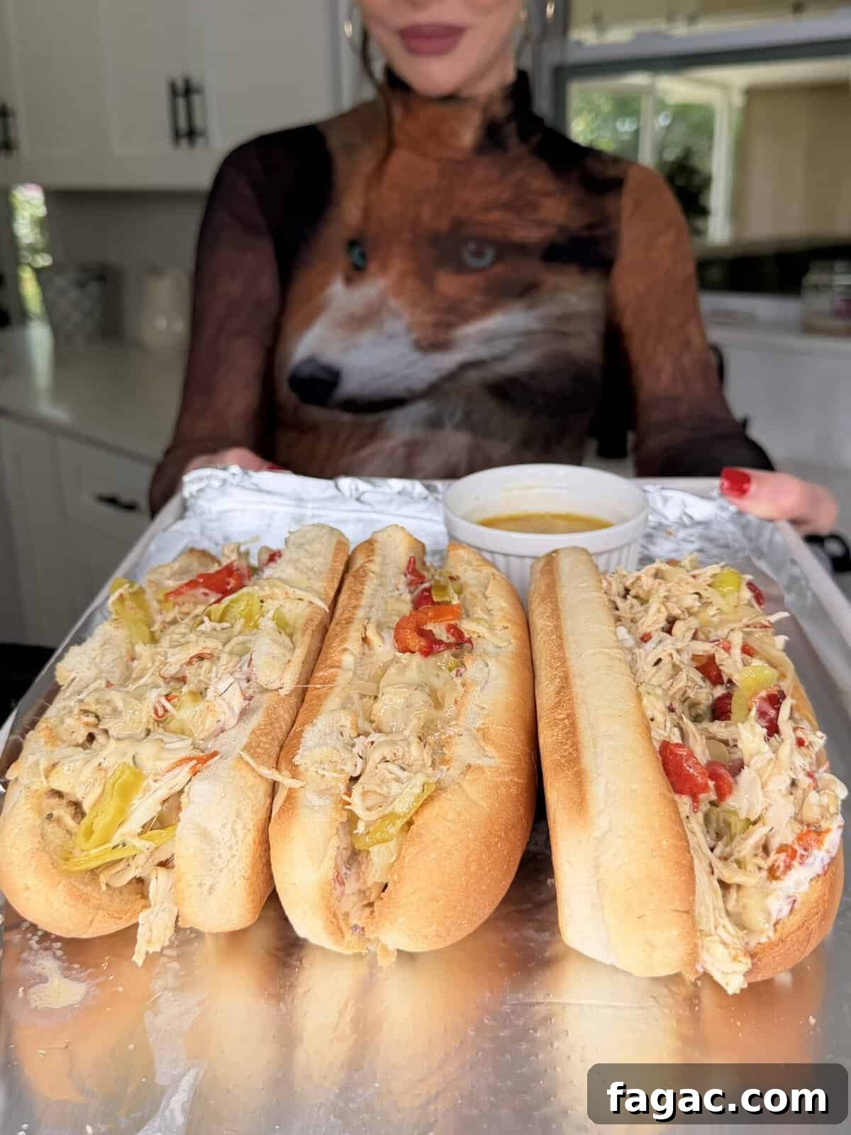 Crockpot Chicken Philly Cheesesteak Recipe for Slow-Cooked Sandwiches 13 A woman in a shirt with a fox design holds a tray of Easy Crockpot Chicken Philly Cheesesteak hoagie rolls filled with shredded chicken, peppers, and cheese, with a small bowl of dipping sauce in a cozy kitchen setting.