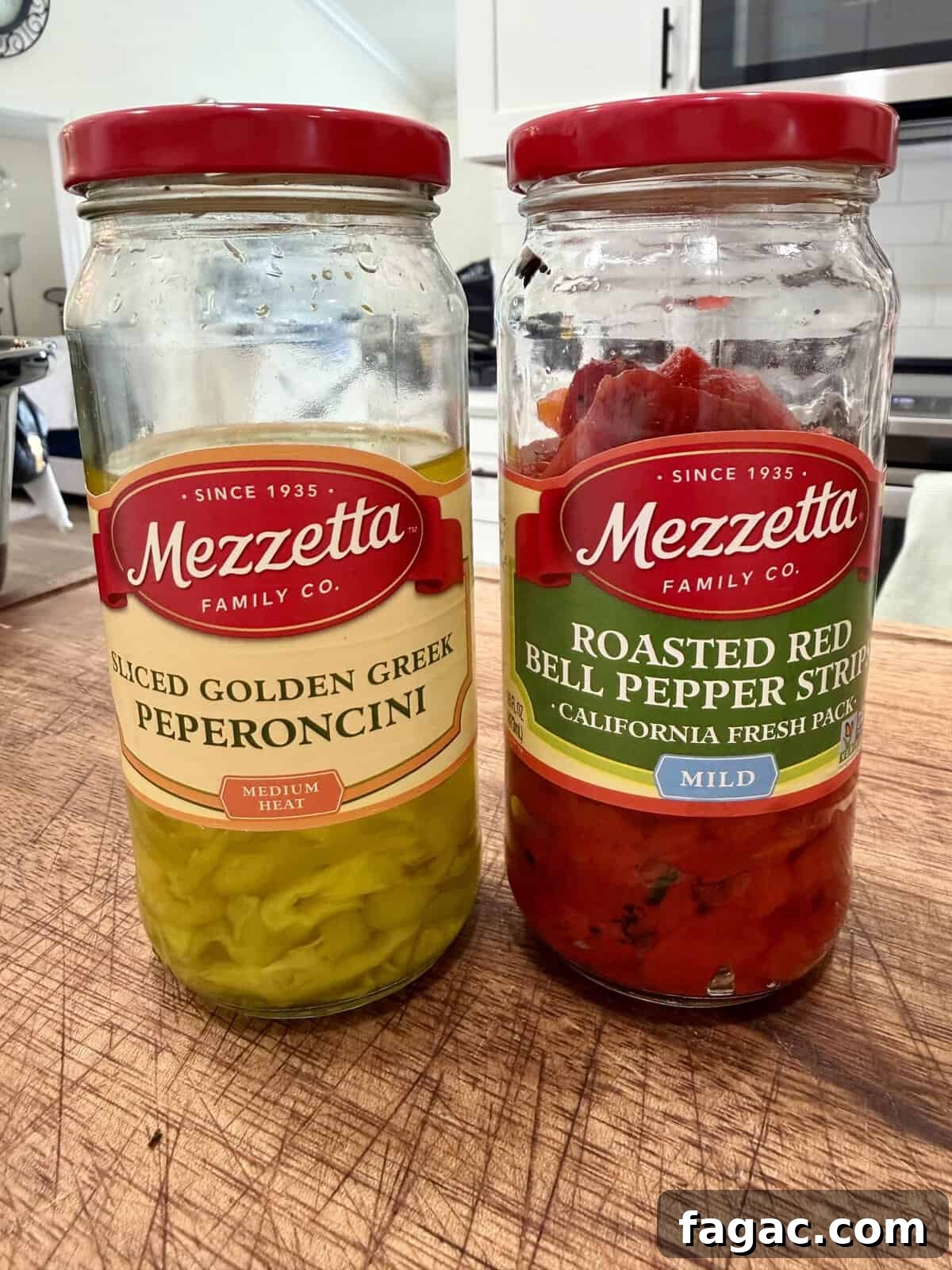 Crockpot Chicken Philly Cheesesteak Recipe for Slow-Cooked Sandwiches 3 Two open jars of Mezzetta brand peppers on a wooden surface: one contains sliced golden Greek pepperoncini, and the other contains roasted red bell pepper strips. A kitchen counter and stove are in the background.