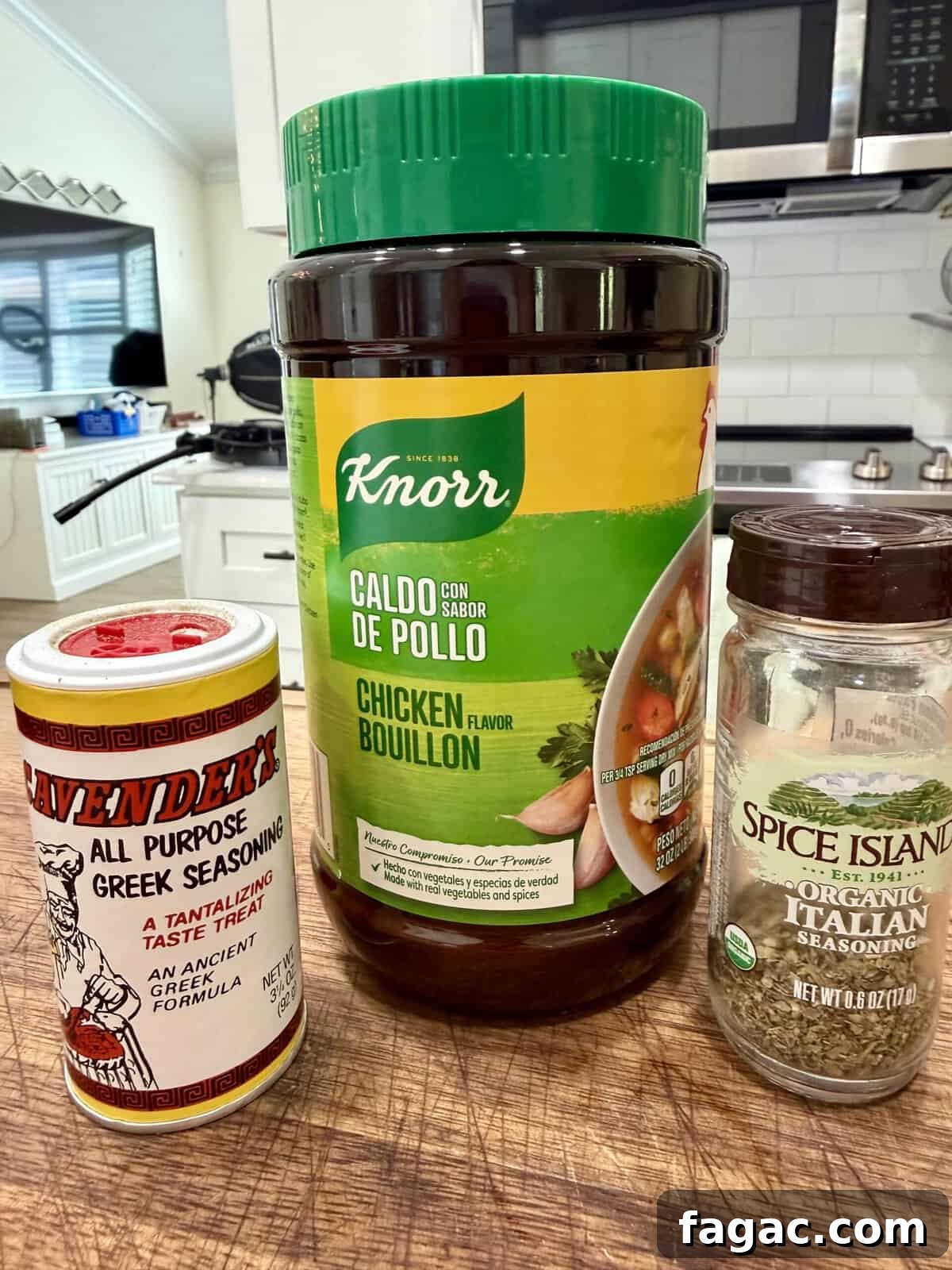 Crockpot Chicken Philly Cheesesteak Recipe for Slow-Cooked Sandwiches 4 Three spice containers—Lawrys Greek Seasoning, Knorr Chicken Bouillon, and Spice Islands Organic Italian Seasoning—stand on a wooden countertop in a kitchen with white cabinets and a stove in the background.