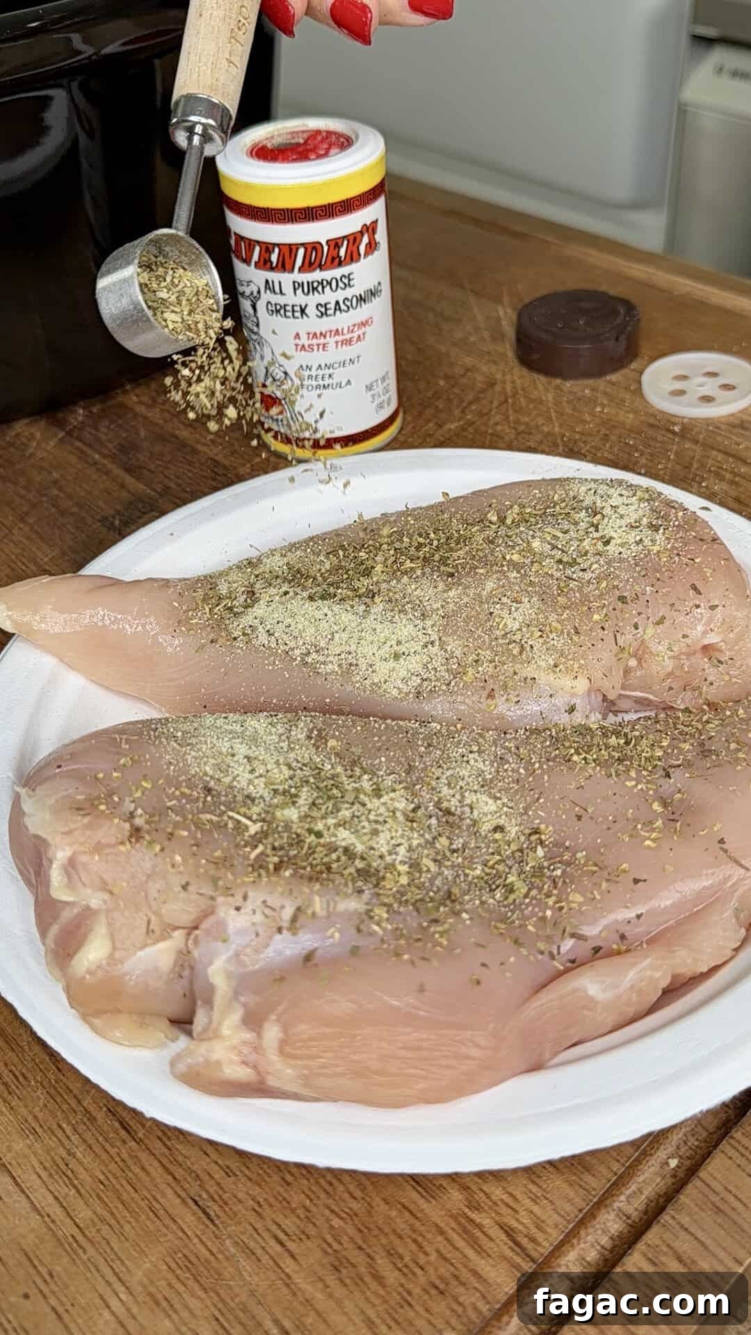 Crockpot Chicken Philly Cheesesteak Recipe for Slow-Cooked Sandwiches 5 A hand sprinkles dried seasoning onto two raw chicken breasts on a white plate. A container of Greek seasoning sits nearby on a wooden surface.