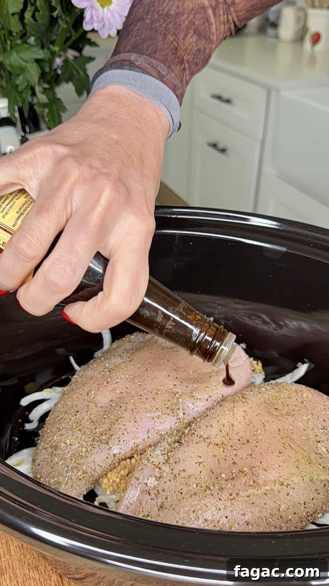 Crockpot Chicken Philly Cheesesteak Recipe for Slow-Cooked Sandwiches 6 A hand pours a dark liquid from a bottle onto seasoned raw turkey breasts placed over sliced onions in a black slow cooker, with a kitchen counter and cabinets in the background.