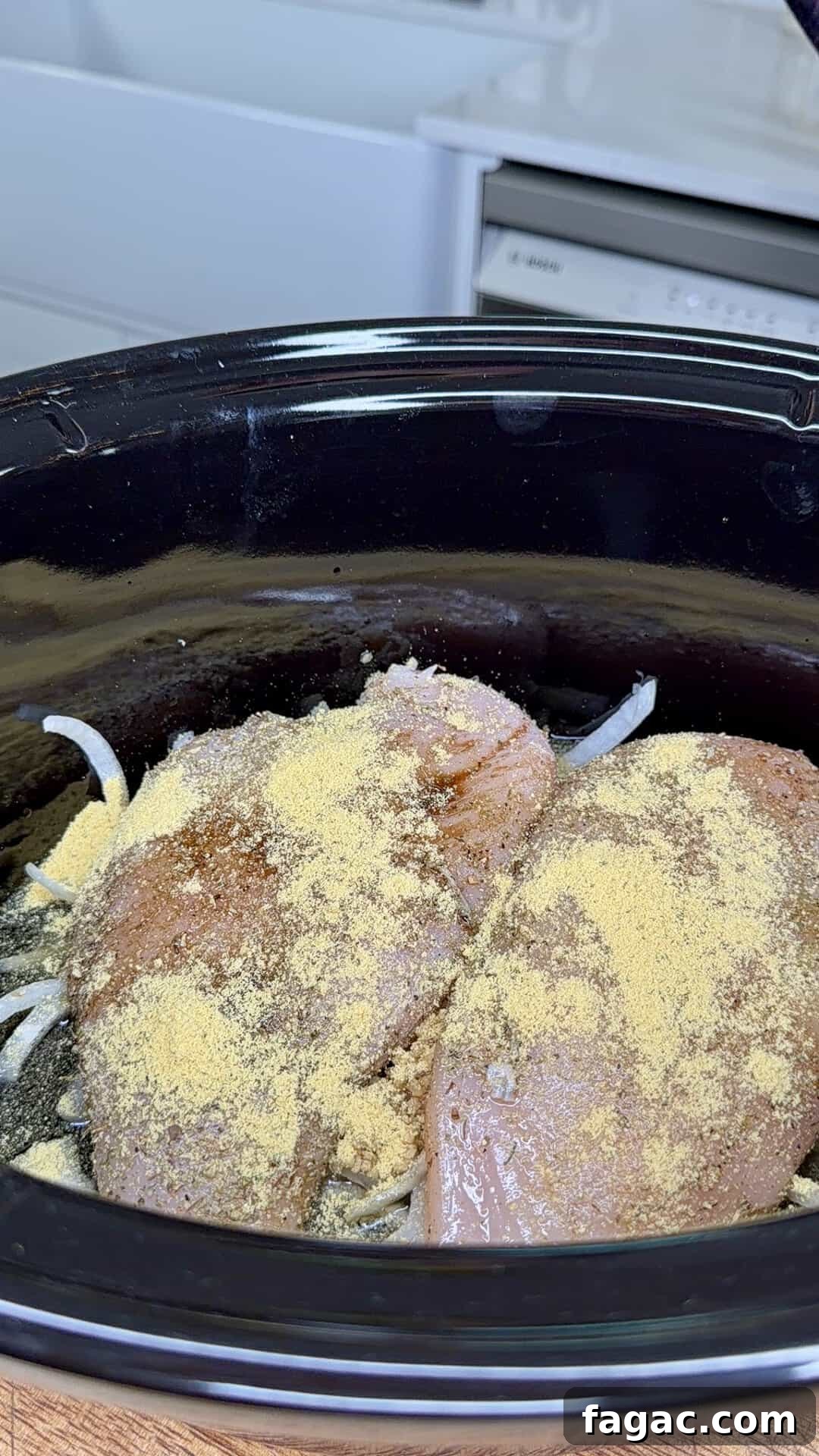 Crockpot Chicken Philly Cheesesteak Recipe for Slow-Cooked Sandwiches 7 Raw chicken breasts covered in seasoning and placed on top of sliced onions inside a black slow cooker, ready to be cooked. Kitchen counters and a dishwasher are visible in the background.