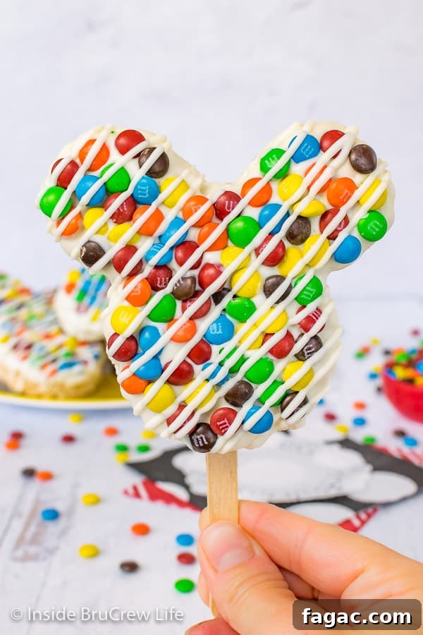 M&M's Mickey Mouse Rice Krispie Treats - these Mickey Mouse shaped treats are one of the easiest Disney copycat recipes to make. Make these Mickey shaped treats and watch Disney fans smile. A close up picture of a M&M's Mickey Mouse Rice Krispie Treat on a stick held up in front of a plate of treats