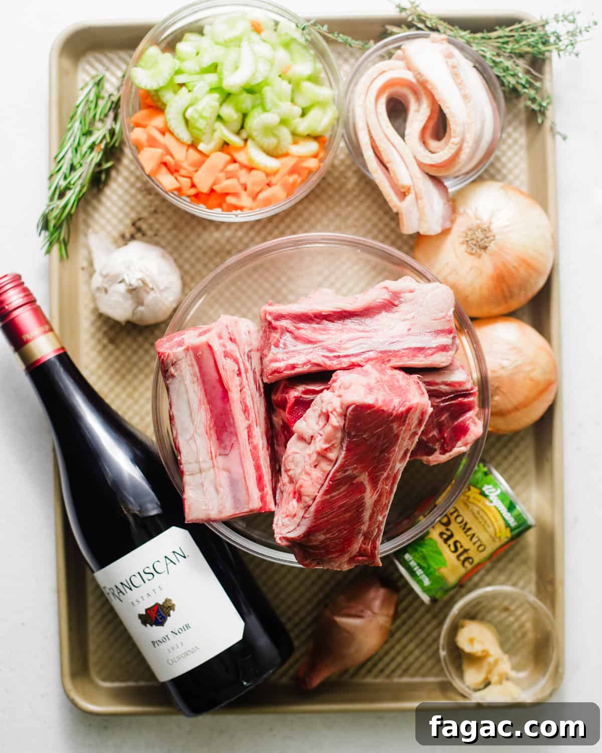 ingredients to make braised short ribs on a baking sheet - short ribs, onion, shallots, bacon, celery, carrots, garlic tomato paste, miso paste, red wine, heavy cream, balsamic vinegar, nutmeg, red pepper flakes, brown sugar, fresh sage, fresh thyme, fresh rosemary, bay leaves, and beef broth.