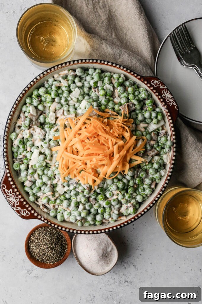 a bowl of easy pea salad with shredded cheddar cheese sprinkled on top next to a little wooden bowl of pepper and a little ceramic bowl of salt