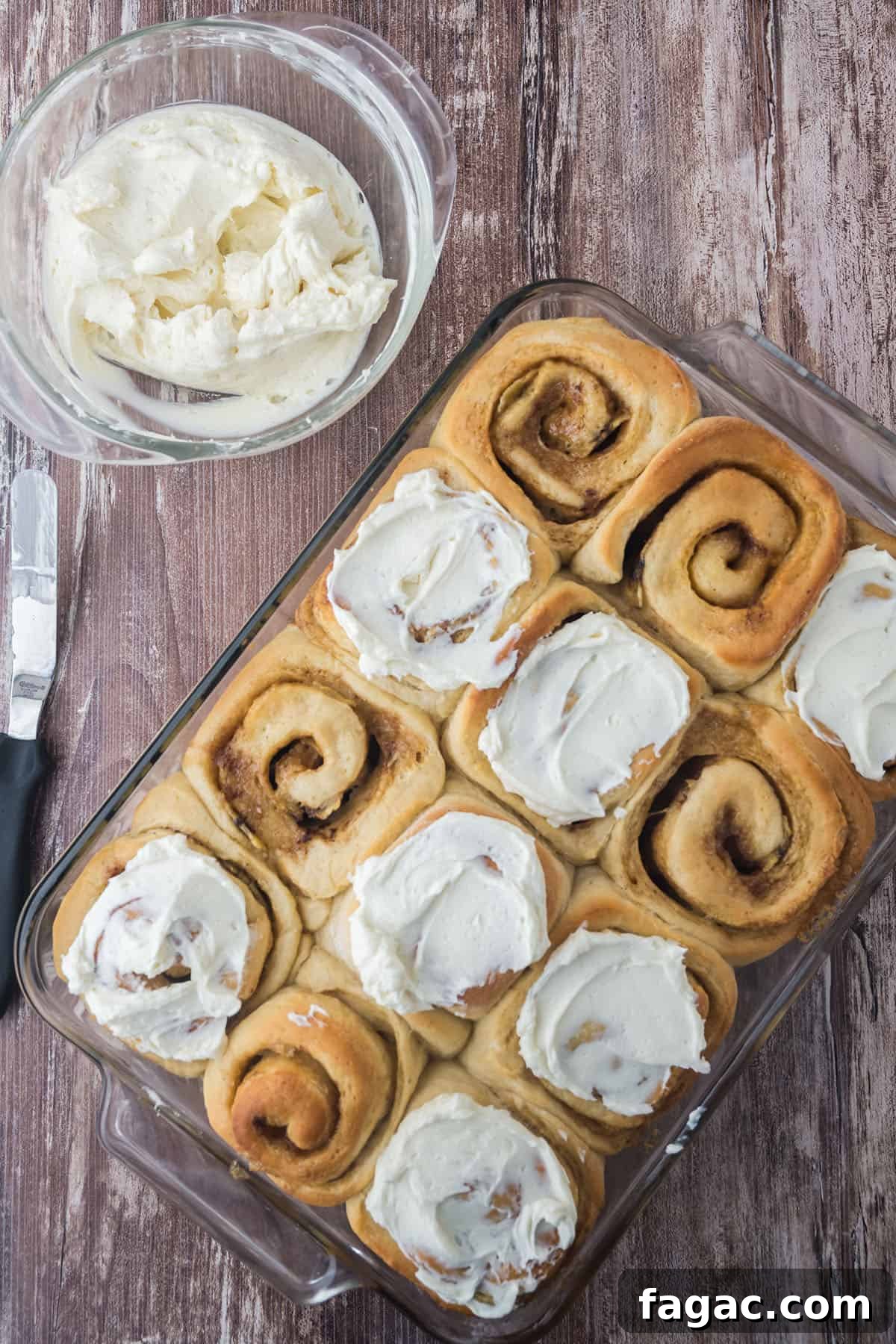 Twelve baked cinnamon rolls in a glass baking dish.  Seven rolls are frosted with cream cheese frosting. A bowl of frosting and a frosting spatula are on the side.