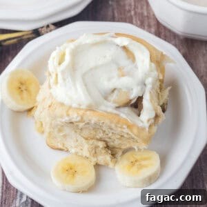 One frosted cinnamon roll on a white plate with some fresh sliced banana on the side.
