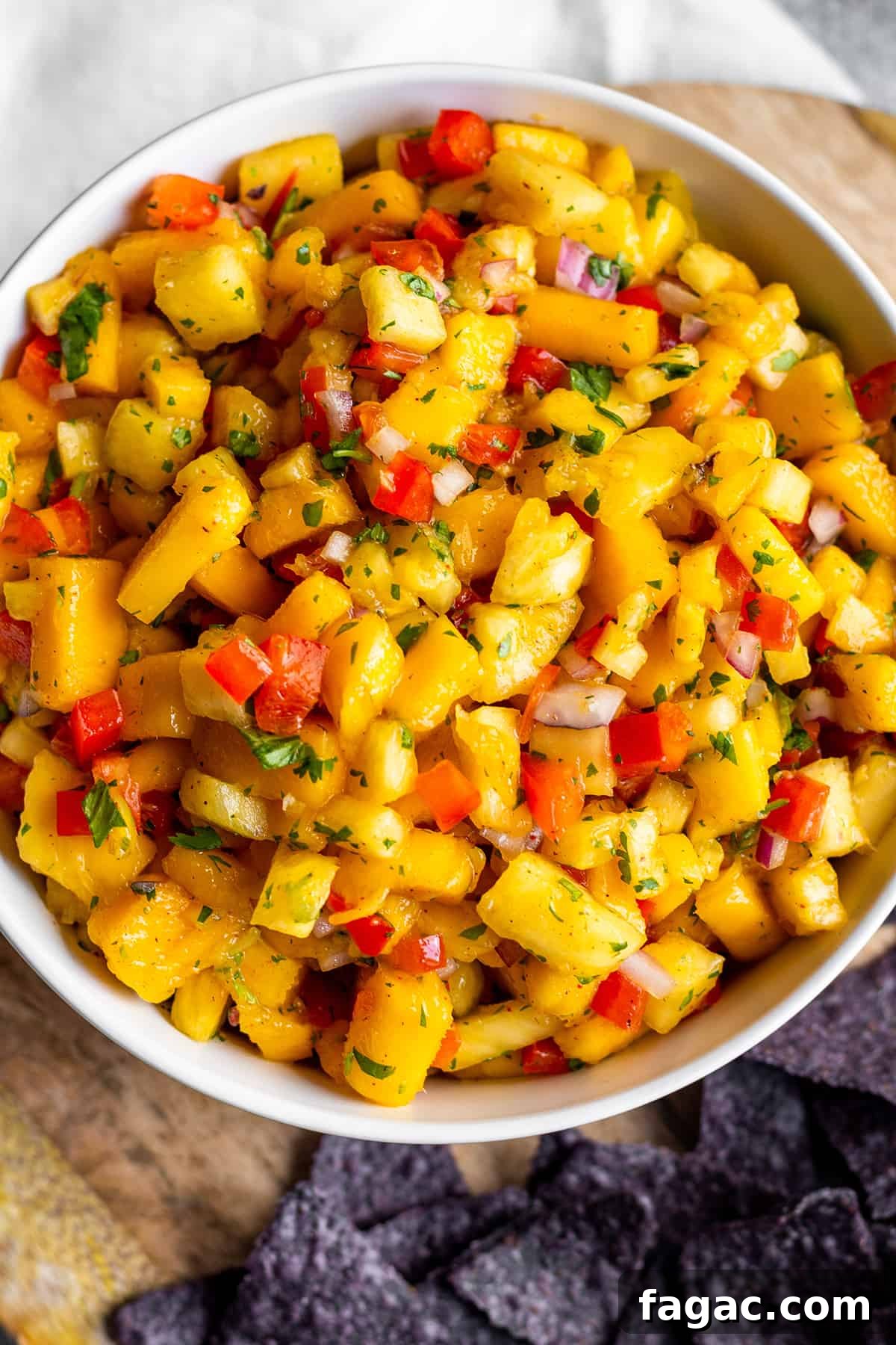 Zesty Mango and Pineapple Salsa Recipe for Summer Parties 5 White bowl with mango pineapple salsa on a wooden board with blue corn tortilla chips.