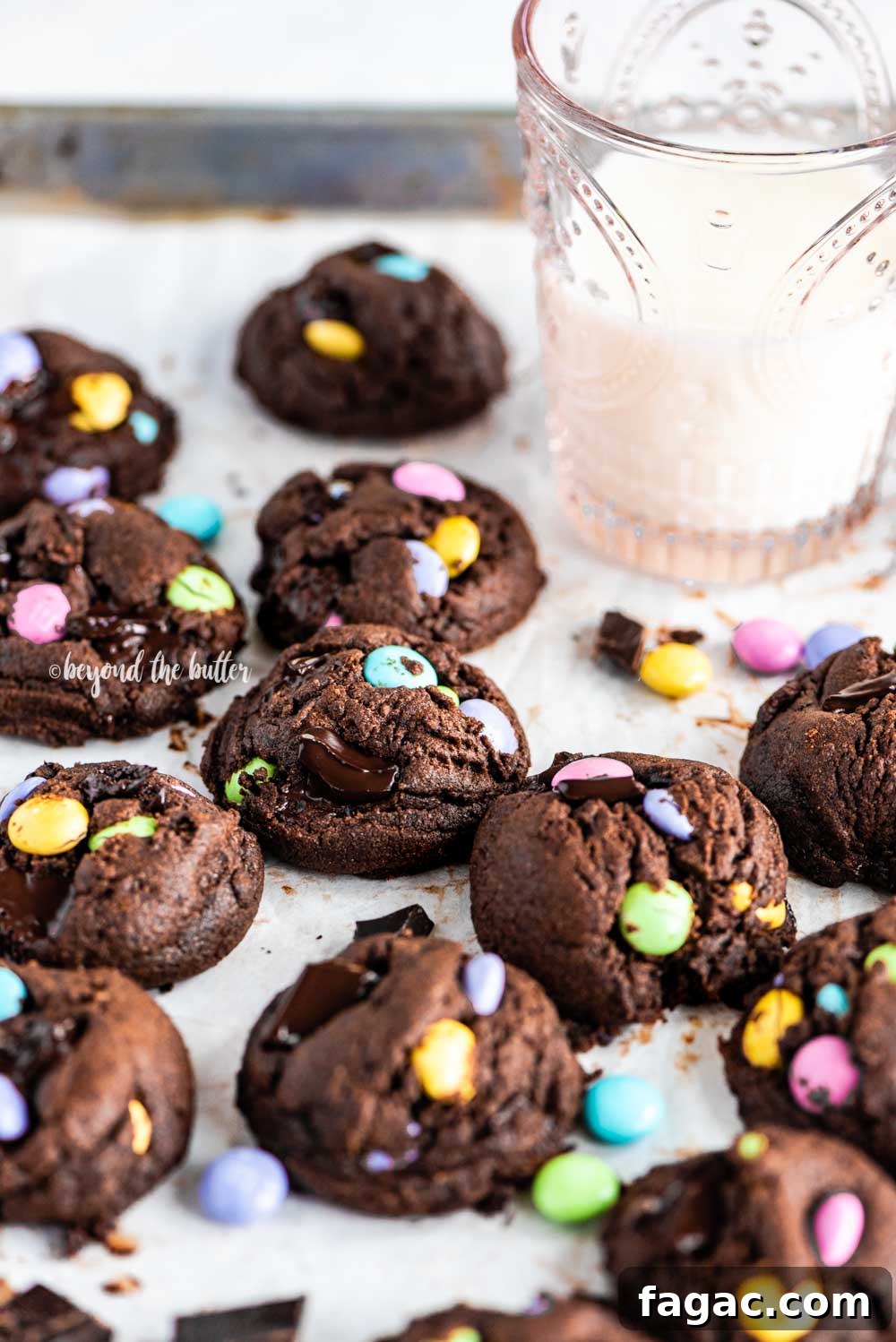Angled image of just baked double chocolate chunk m&m cookies on a parchment lined baking sheet and a glass of milk above it | All Images © Beyond the Butter™