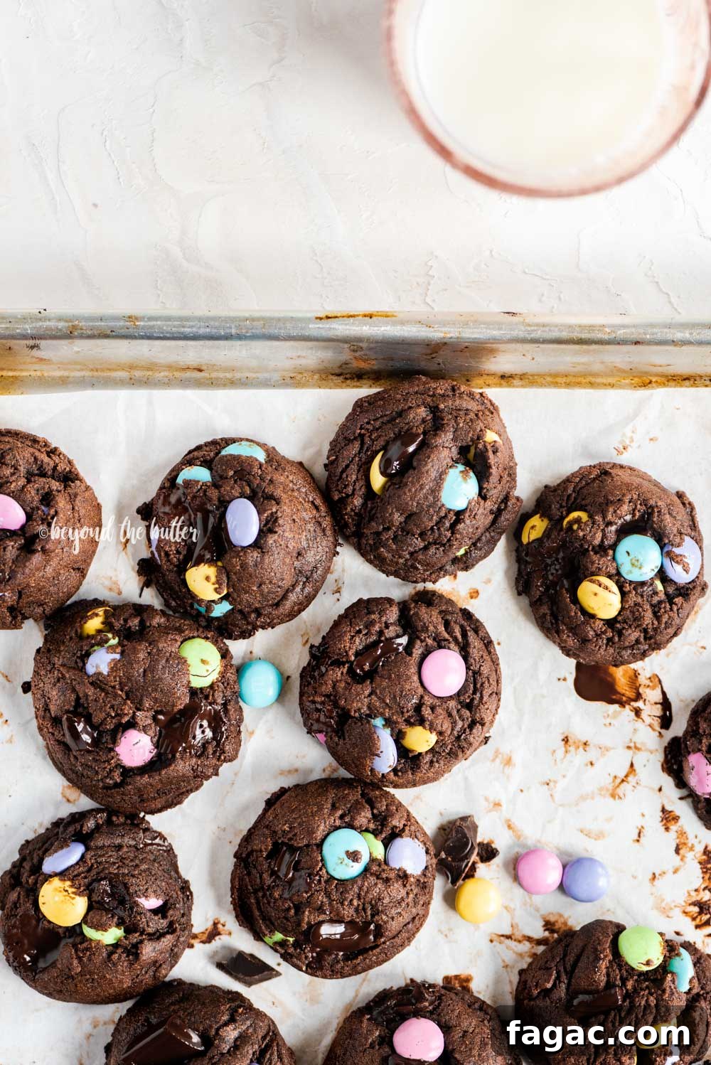 Overhead image of just baked double chocolate chunk m&m cookies on a parchment lined baking sheet and a glass of milk above it | All Images © Beyond the Butter™