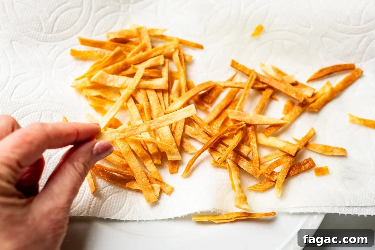Quick Chicken Tortilla Soup Recipe for Cozy Weeknight Meals 16 fingers holding a fried tortilla strip, with a pile of fried strips on a paper towel behind it.