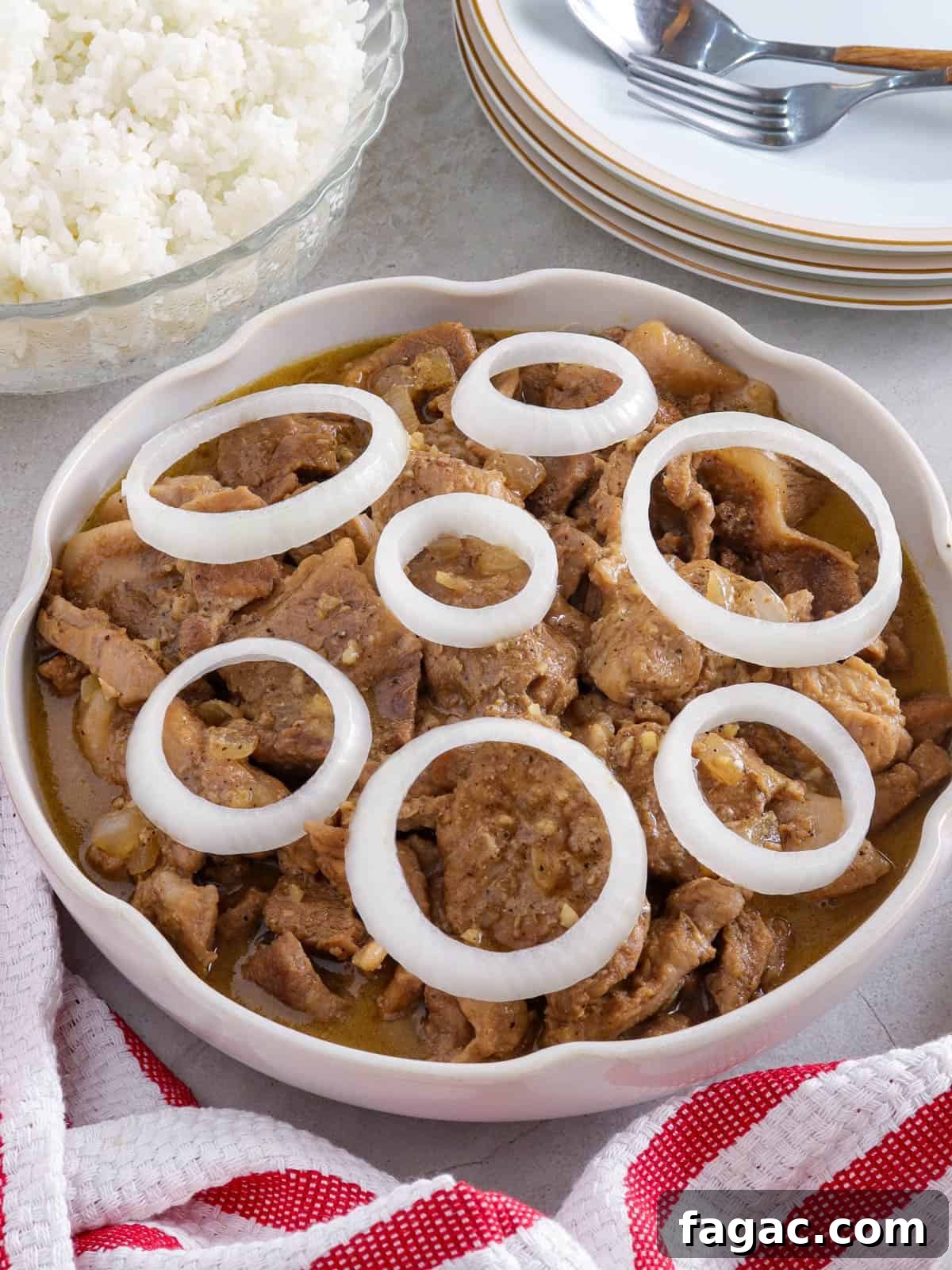 Creamy Pork Steak with onion rings in a white serving bowl with a bowl of steamed rice in the background.