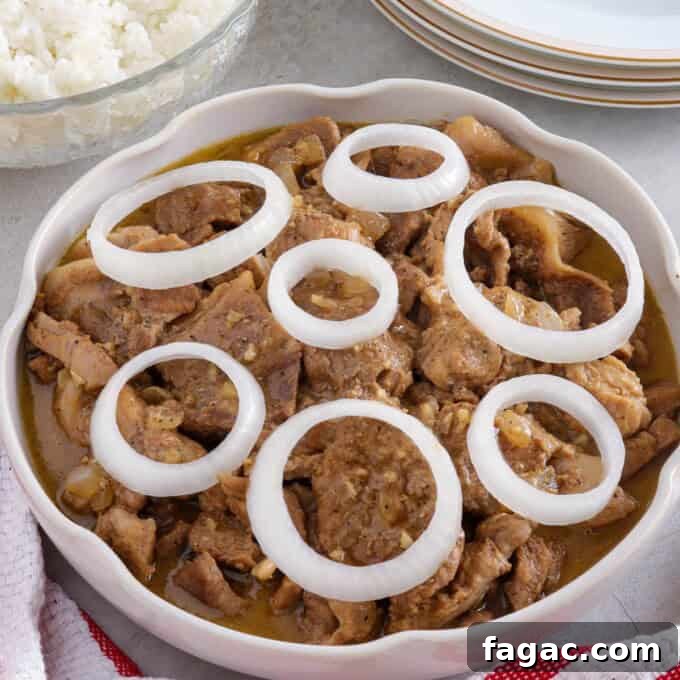 Creamy Pork Steak with onion rings in a white serving bowl with a bowl of steamed rice in the background.