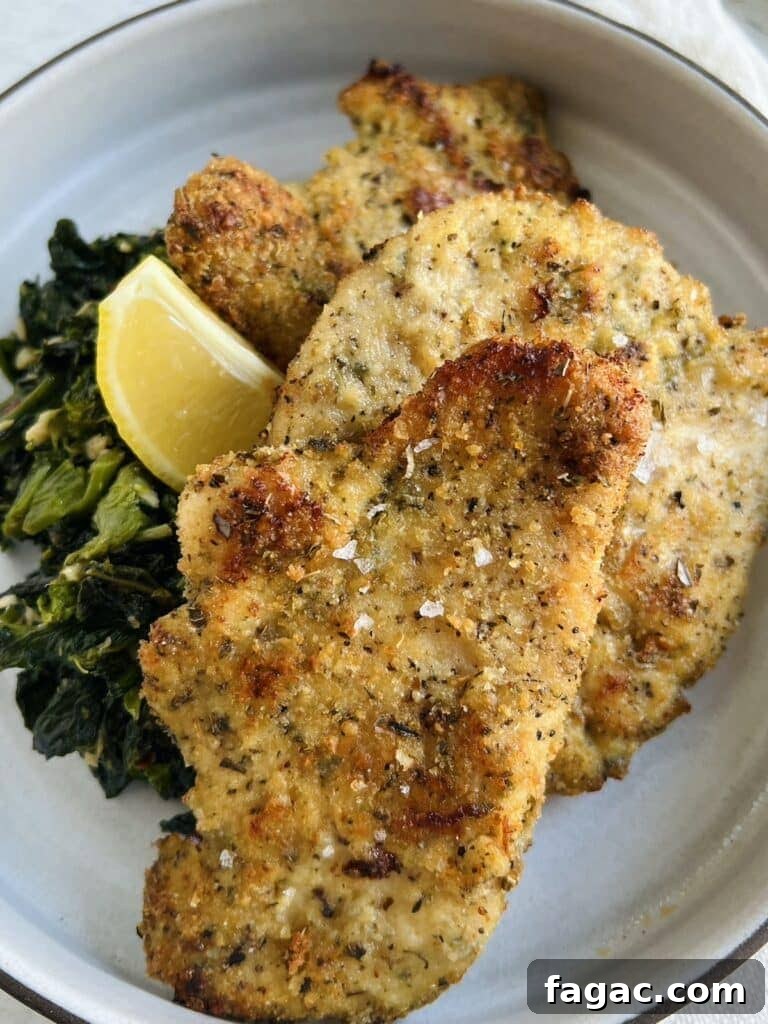 Baked chicken cutlets, Italian style, on plate with sautéed broccoli rabe and lemon wedge.