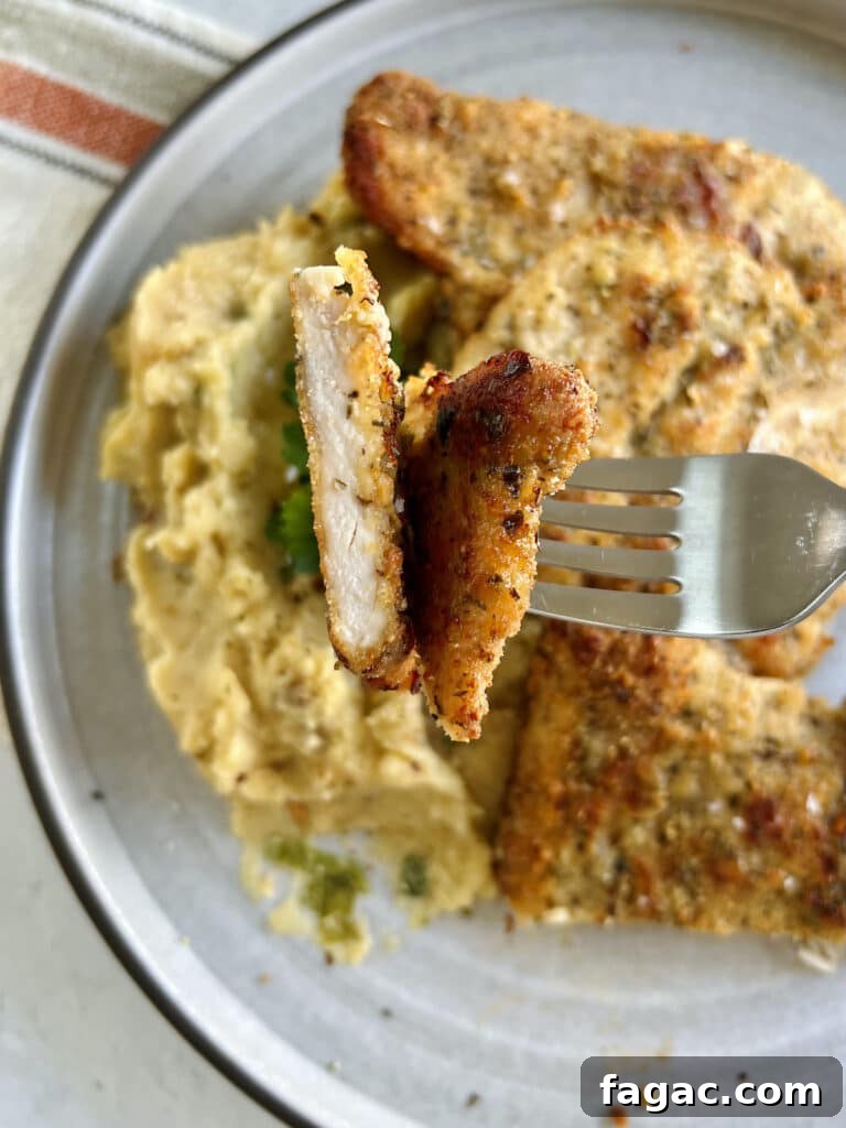 Close up of piece of baked chicken cutlet on fork held over plate of cutlets and mashed potatoes.