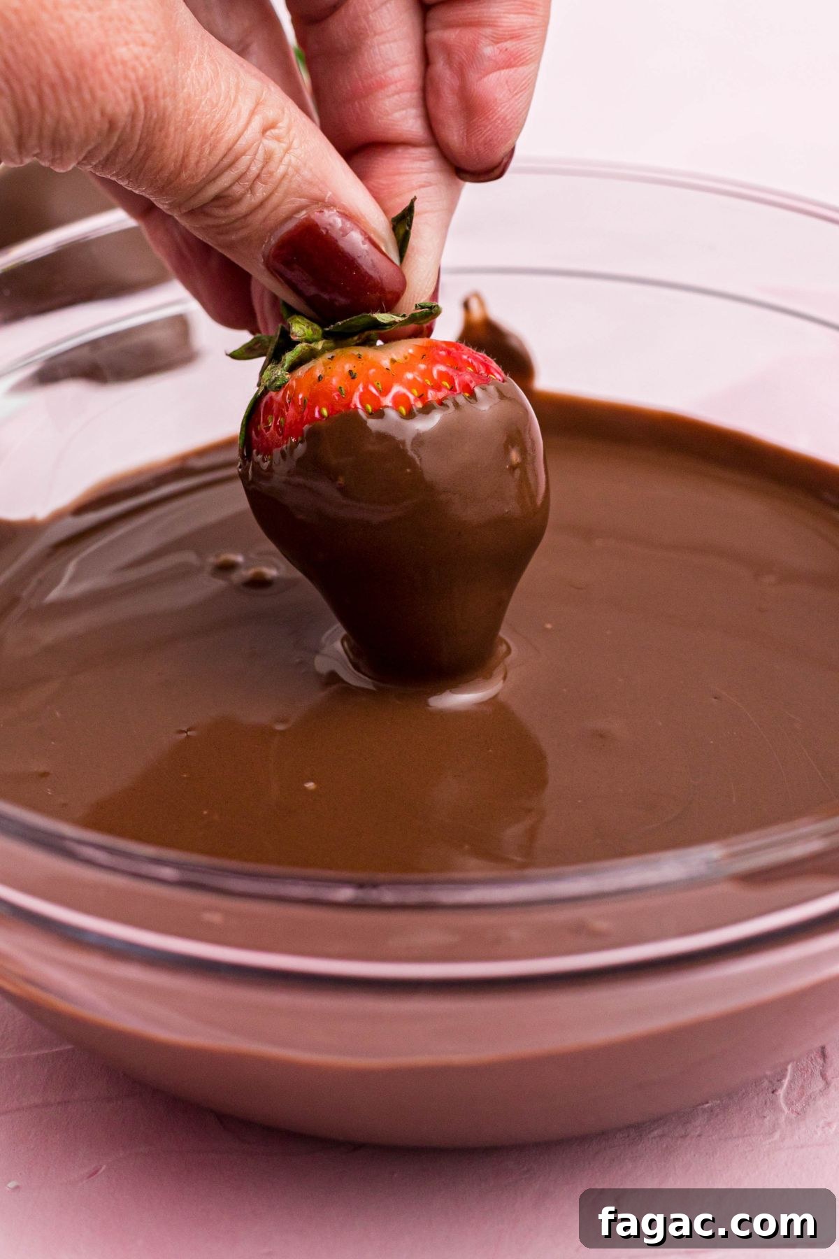 How to Make Chocolate-Covered Strawberries 4 Fresh strawberry being dipped into melted milk chocolate in a clear glass bowl.